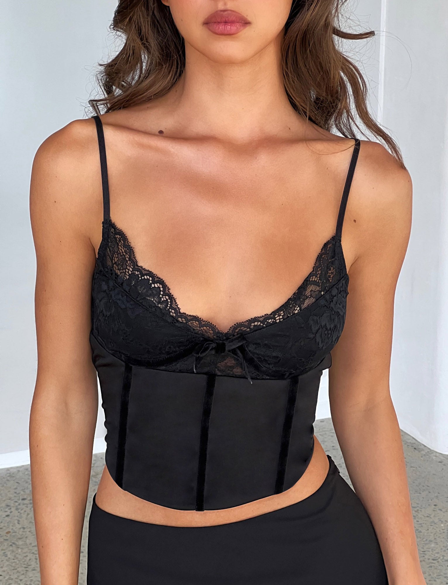 A woman is wearing the MORENA TOP - BLACK from TIGERMIST, a black lace-trimmed camisole with spaghetti straps and a bow detail in the center. Her midriff is visible as she poses against a plain, neutral-colored background.