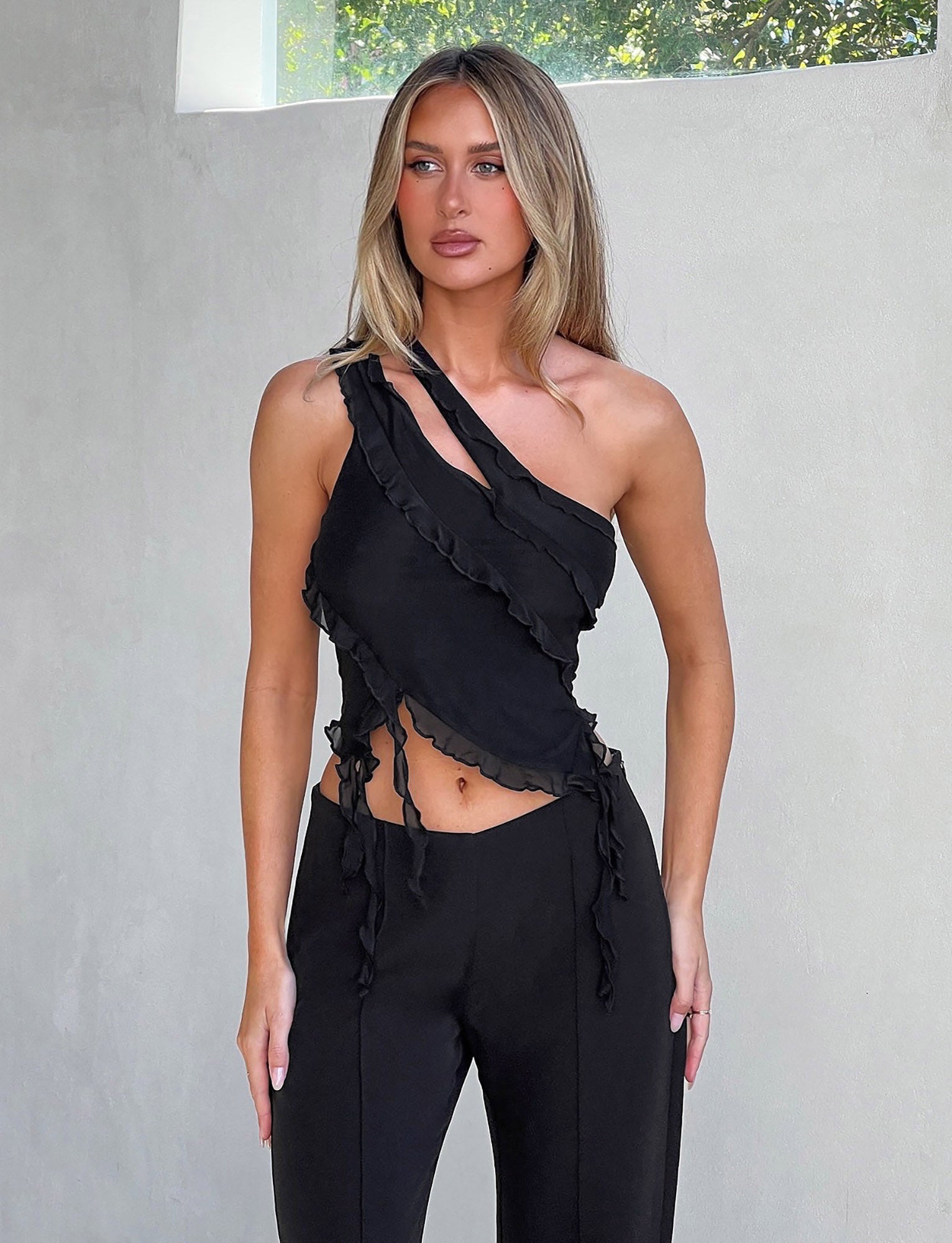 A woman with long blonde hair is standing against a light gray wall, wearing the stylish DIONNE TOP - BLACK from TIGERMIST and fitted black pants. The top features an asymmetrical design with ruffled edges, revealing parts of her shoulders and midriff. Natural light illuminates her.