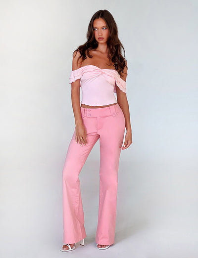 A woman stands against a plain backdrop wearing the DENALI TOP - PINK and matching pink high-waisted pants from TIGERMIST. She has long brown hair and wears white open-toe high heels. Her left hand rests on her hip, and she gazes directly at the camera.