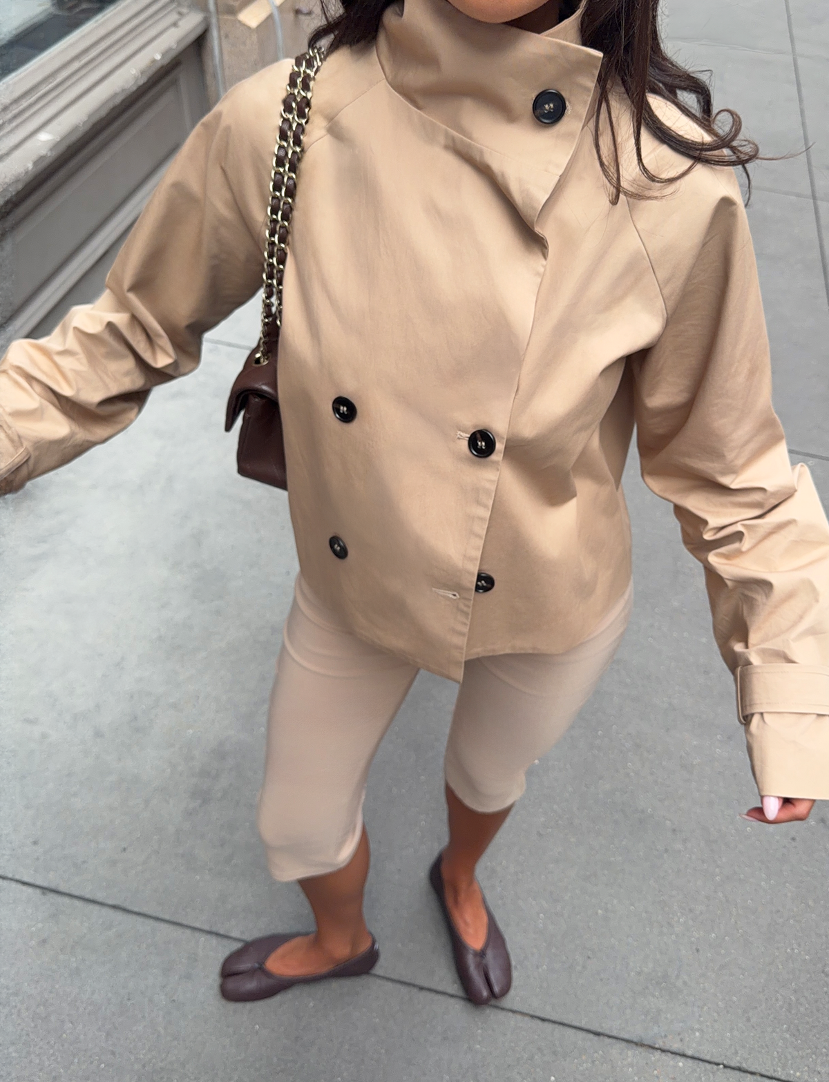 A woman stands on a city sidewalk, wearing the TIGERMIST HAYLEY CROPPED TRENCH JACKET in khaki, paired with beige pants and brown flats. She holds a brown chain-strap handbag, captured from above with her face out of view.
