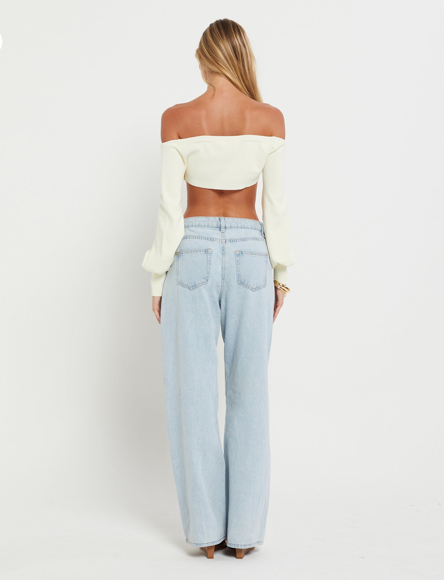 A woman stands facing away from the camera, showcasing her off-the-shoulder long-sleeve MARIYAH Jumper in cream by TIGERMIST and light blue high-waisted jeans. She has long, blonde hair that flows down her back and is standing against a plain white background.