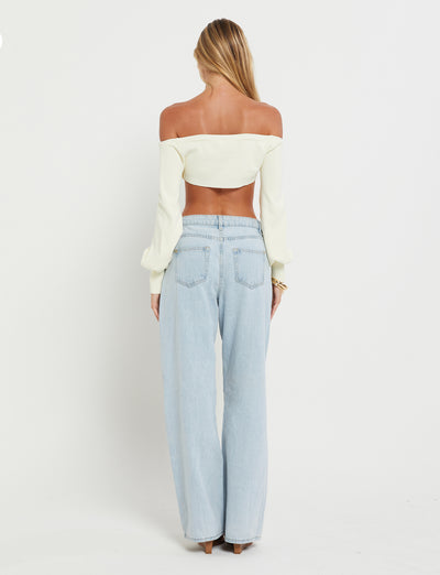 A woman stands facing away from the camera, showcasing her off-the-shoulder long-sleeve MARIYAH Jumper in cream by TIGERMIST and light blue high-waisted jeans. She has long, blonde hair that flows down her back and is standing against a plain white background.