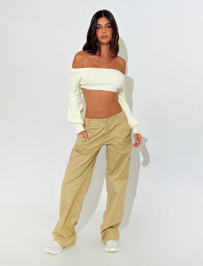 A woman stands against a plain background, wearing the MARIYAH JUMPER - NEUTRAL in cream from TIGERMIST paired with beige cargo pants. She has long, wavy hair and is posing with her hands in her pants pockets. She completes the look with white sneakers.