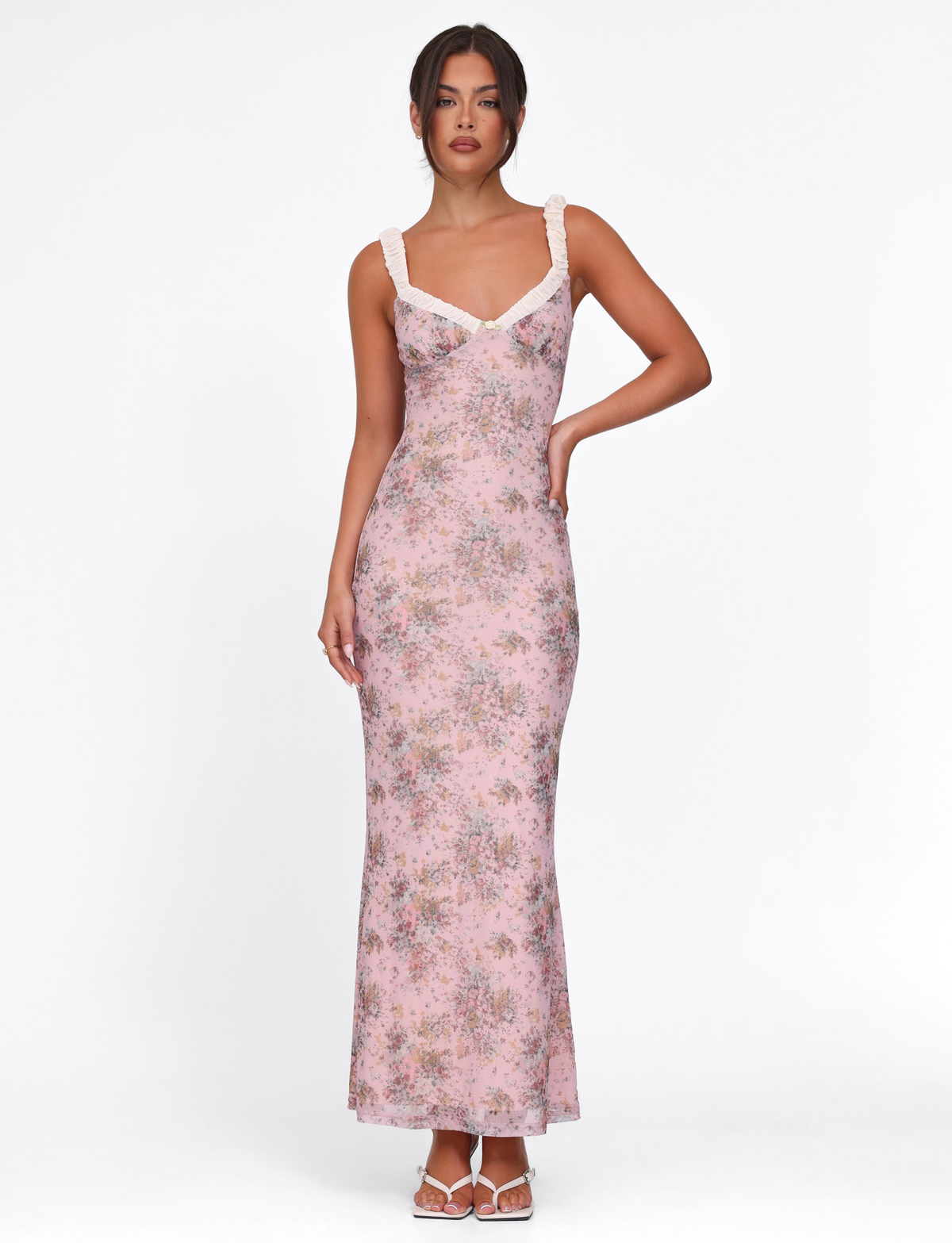 A woman poses against a white background wearing the TIGERMIST AMARAN DRESS - PINK : CAMO FLORAL, featuring ruffled straps and a bodycon silhouette. Her hair is up, one arm at her waist, paired with white sandals.
