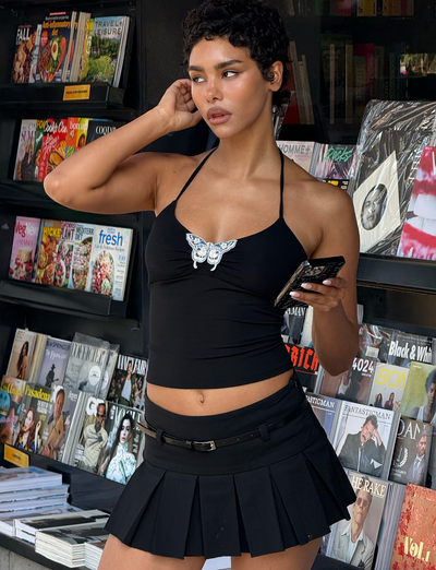 Wearing the TIGERMIST ANNELISE SKORT - BLACK, a person in a black butterfly halter top stands by a magazine stand, holding a phone to their ear and looking to the side with magazines displayed behind them.