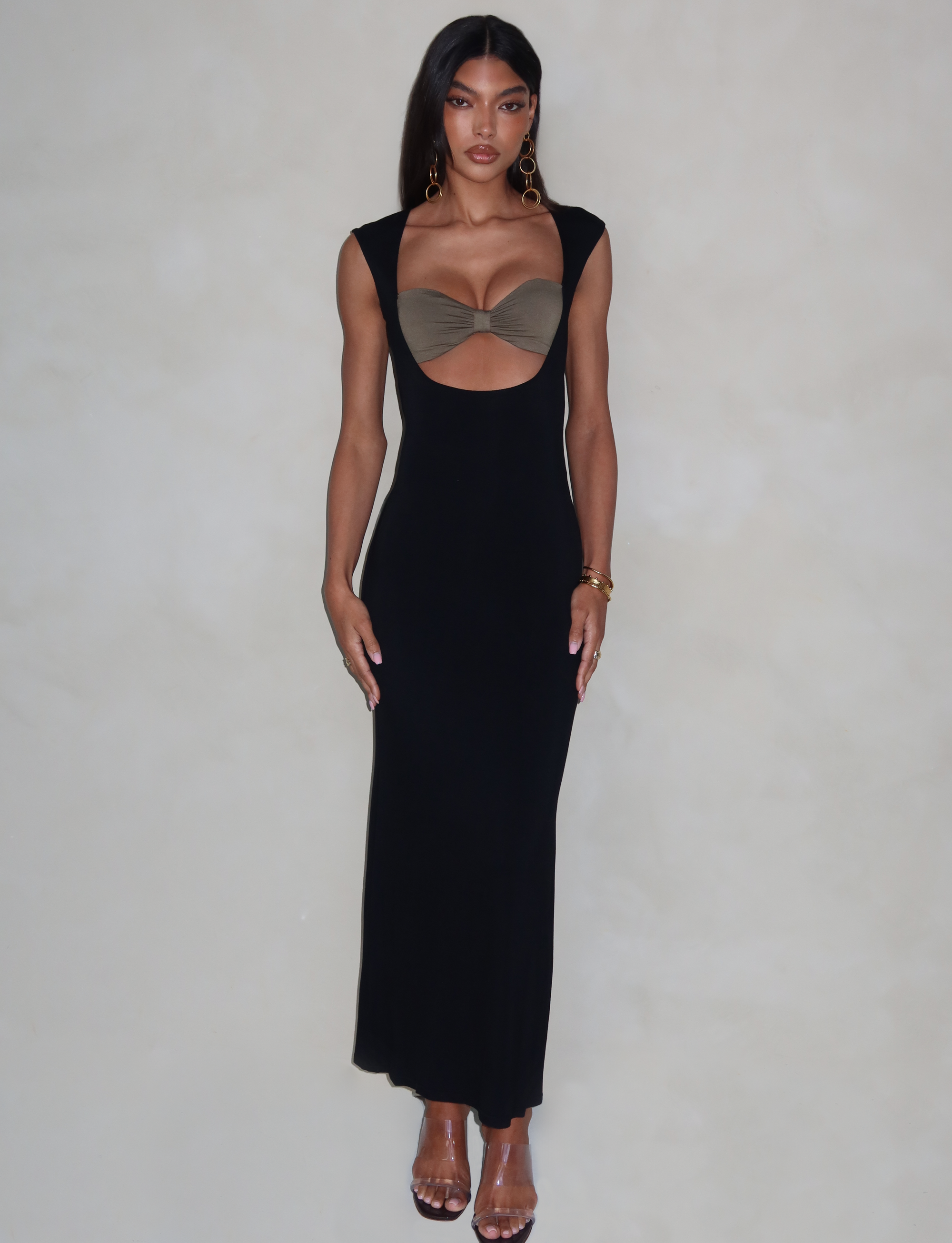 A woman poses stylishly against a light background in a fitted silhouette, wearing the TIGERMIST BLAKELY DRESS in black, featuring a front cutout with brown details. She completes her ensemble with gold hoop earrings, bracelets, and clear high-heeled shoes.