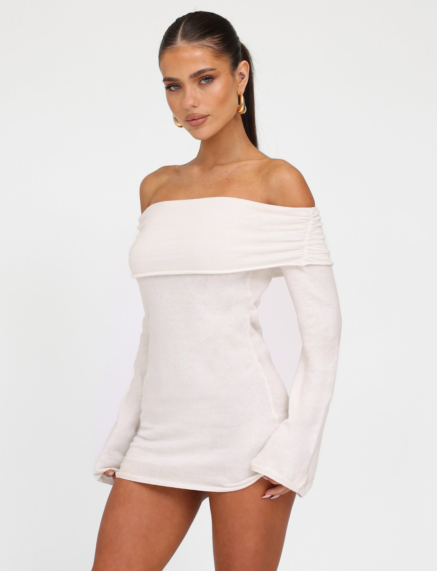 A woman with long dark hair is wearing the DIEDRE JUMPER DRESS - WHITE : CREAM from TIGERMIST. She stands confidently with her hand on her hip, looking directly at the camera. She accessorizes with large gold hoop earrings against a plain white background.