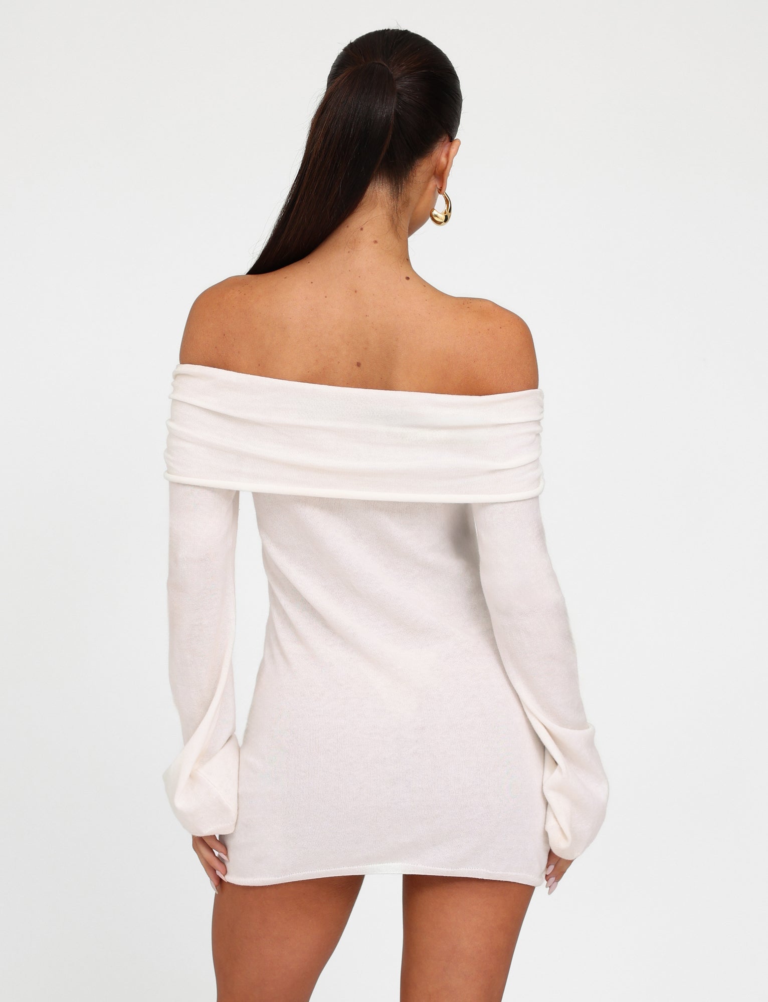 A woman with long dark hair tied in a ponytail is standing with her back to the camera. She is wearing an off-shoulder, long-sleeved, short Diedre Jumper Dress - White : Cream from TIGERMIST and gold hoop earrings. The dress drapes elegantly over her shoulders, exposing her upper back.