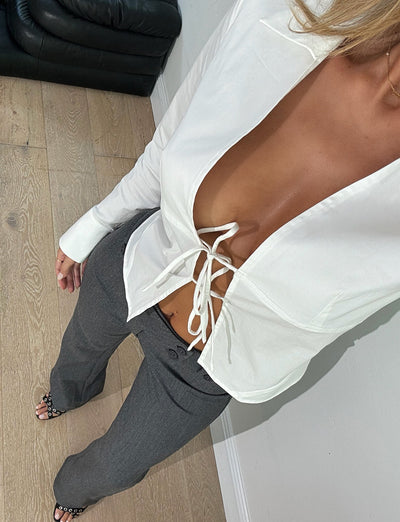 A person is standing on a wooden floor, wearing the ELVA SHIRT - WHITE from TIGERMIST tied at the midriff with strings, grey pants, and black heeled sandals. Only parts of the person's torso and legs are visible in the image.
