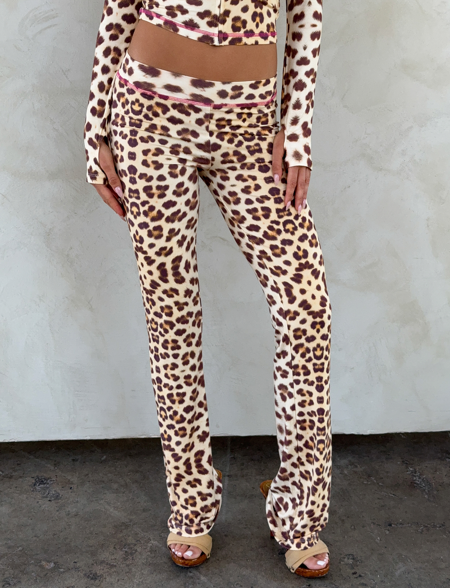 A person stands against a light textured wall, wearing I.AM.GIA's BLARE TRACKPANT in NEUTRAL : LEOPARD : WILD CARD, paired with a matching long-sleeve crop top. Their hands rest near their thighs.