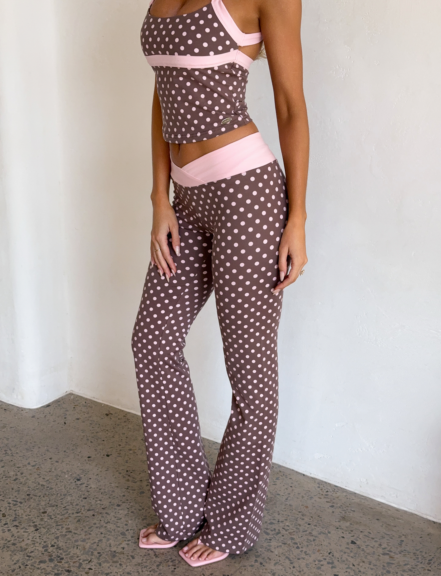 A woman stands indoors in I.AM.GIA's BLARE CROSSOVER PANT - PINK CHOCOLATE with a polka dot print and crossover waistband, paired with a matching tank top and pink sandals. Her face is not visible against a plain white wall.