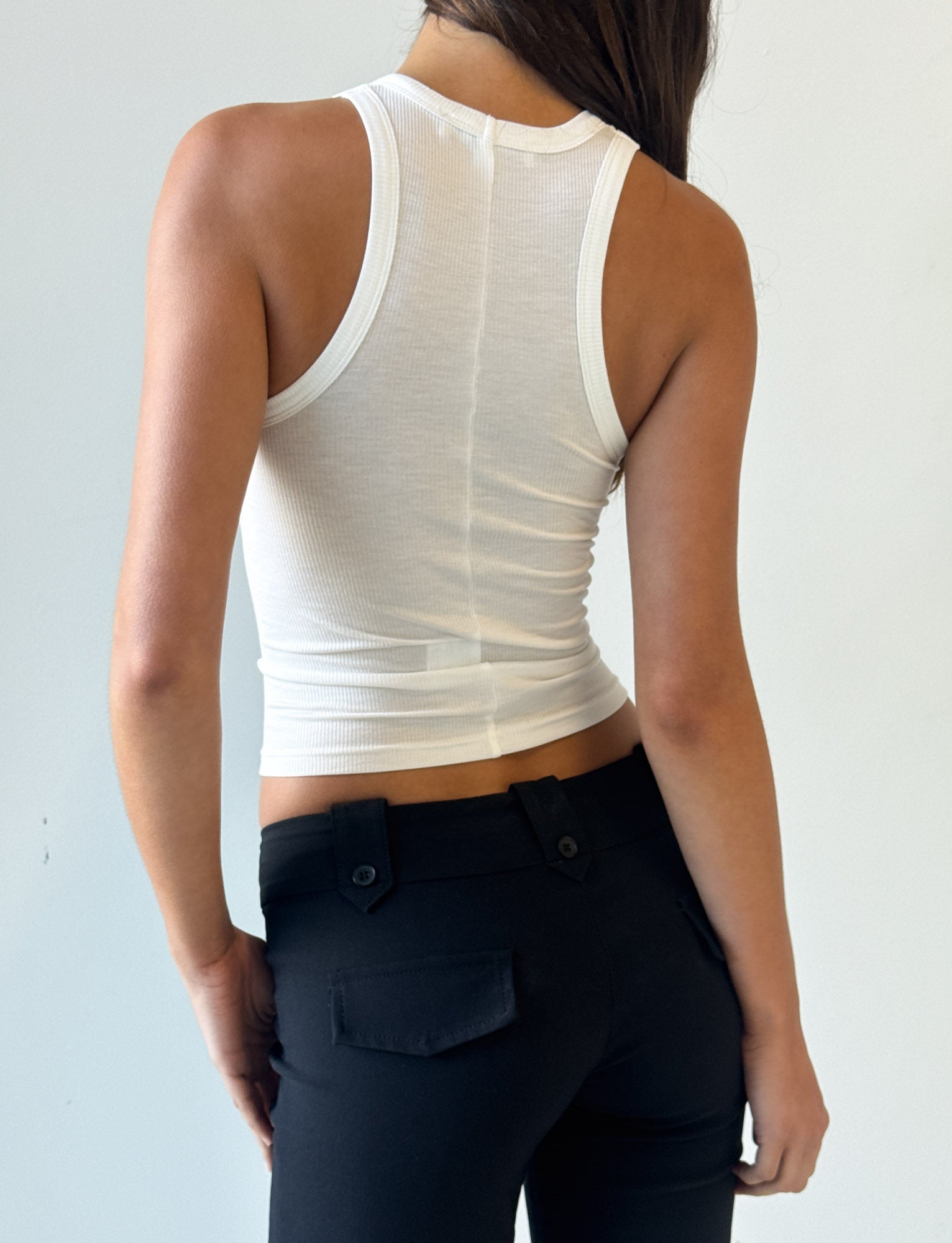A person stands facing away, wearing the I.AM.GIA PENELOPE TANK - PINK AIRBRUSH with ribbed fabrication and black pants with buttoned pockets, set against a plain light background.