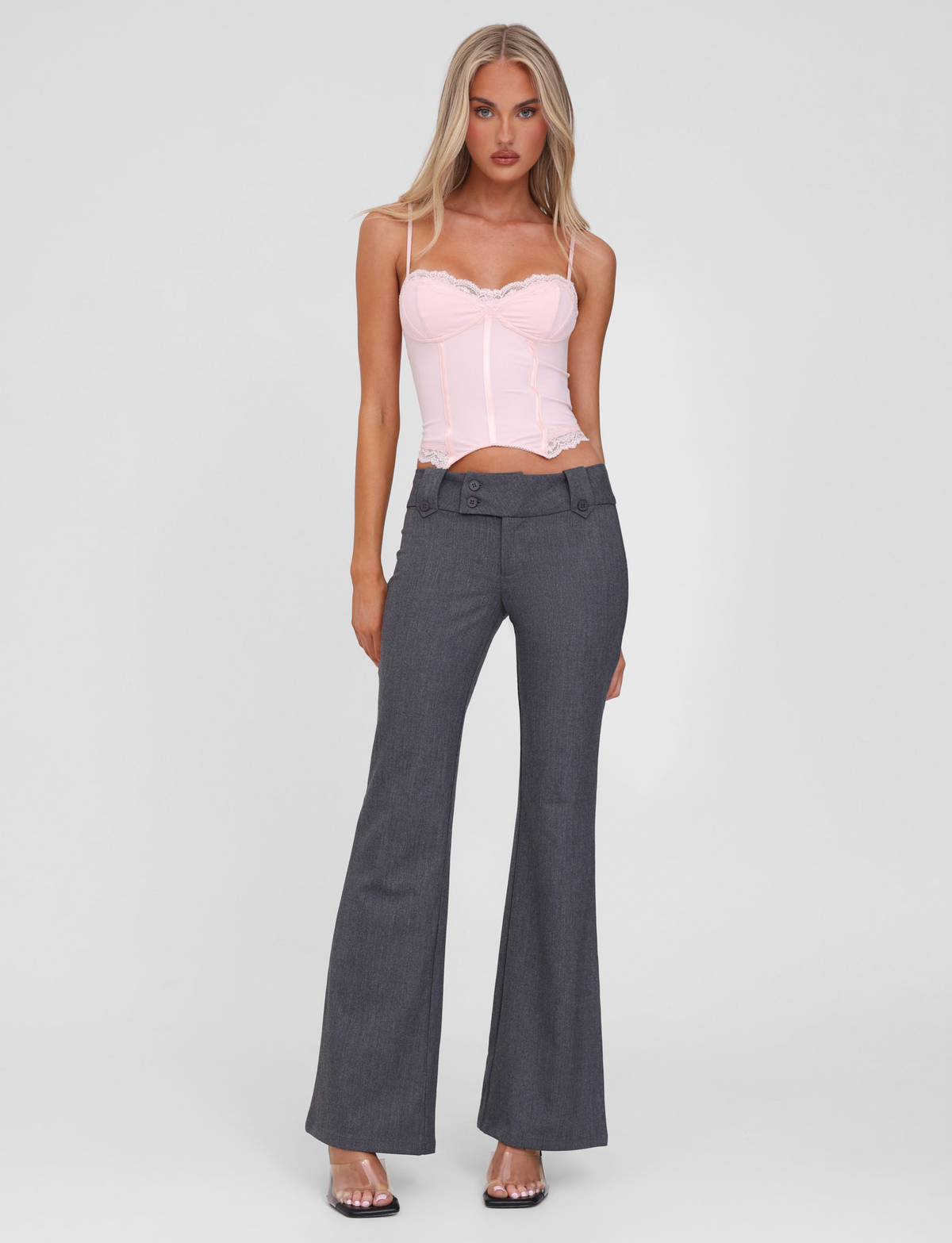 A woman stands against a plain background wearing a light pink bustier-style top and TIGERMIST KITTIE PANT - GREY with flared legs and button detailing at the waistband, paired with clear high-heeled shoes.