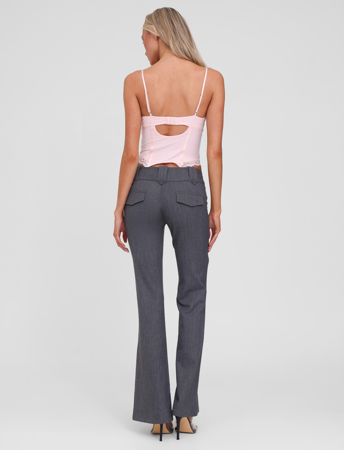 A woman with long blonde hair is seen from the back, wearing a pale pink strappy top and TIGERMIST's KITTIE PANT - GREY with flared legs and flap pockets, paired with black heels against a plain light background.