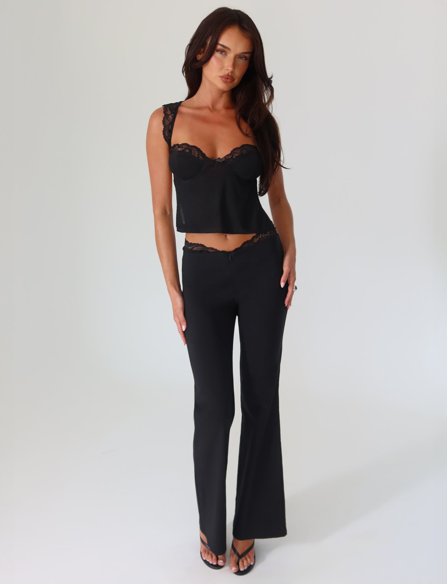 A woman poses confidently in the high stretch fabric from Tigermist, showcasing the stylish NYOMI PANT - BLACK. Her sleeveless top features chic black lace details, and she pairs it perfectly with the flared leg pants. With her long dark hair cascading down, she stands against a plain white background.