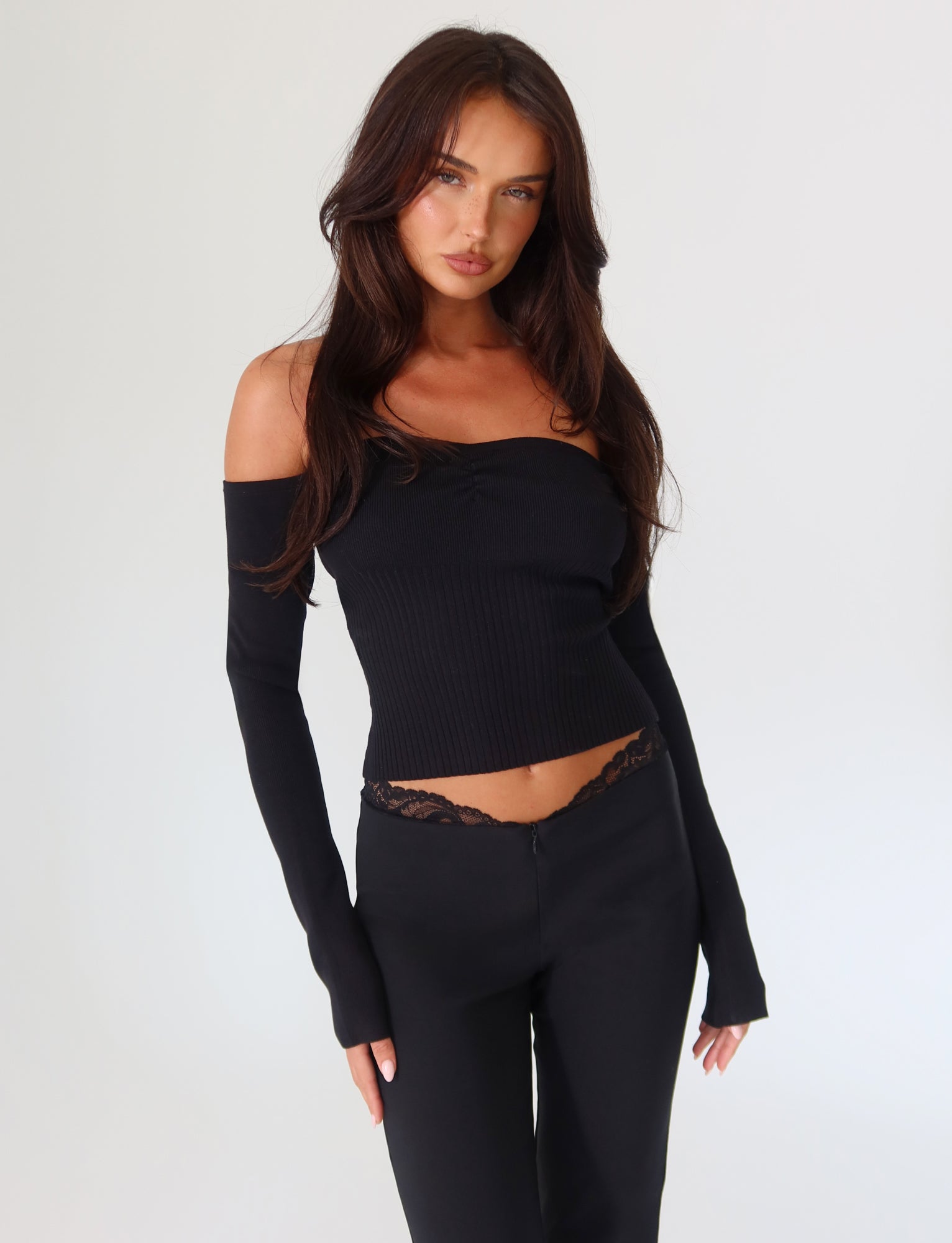 A woman with long brown hair stands against a plain white background, wearing the LILYANNA LONG SLEEVE TOP in black from TIGERMIST, along with black pants featuring lace trim. Her hands are relaxed by her sides.