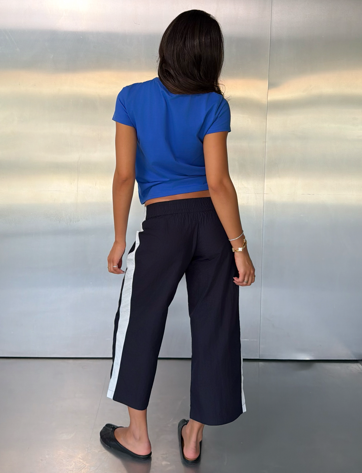 A woman with long dark hair stands facing away, wearing the TIGERMIST SOPHIA WINDBREAKER CAPRI in navy and white—black wide-leg windbreaker pants featuring white side stripes—paired with a blue t-shirt and black flats, in front of a metallic wall.