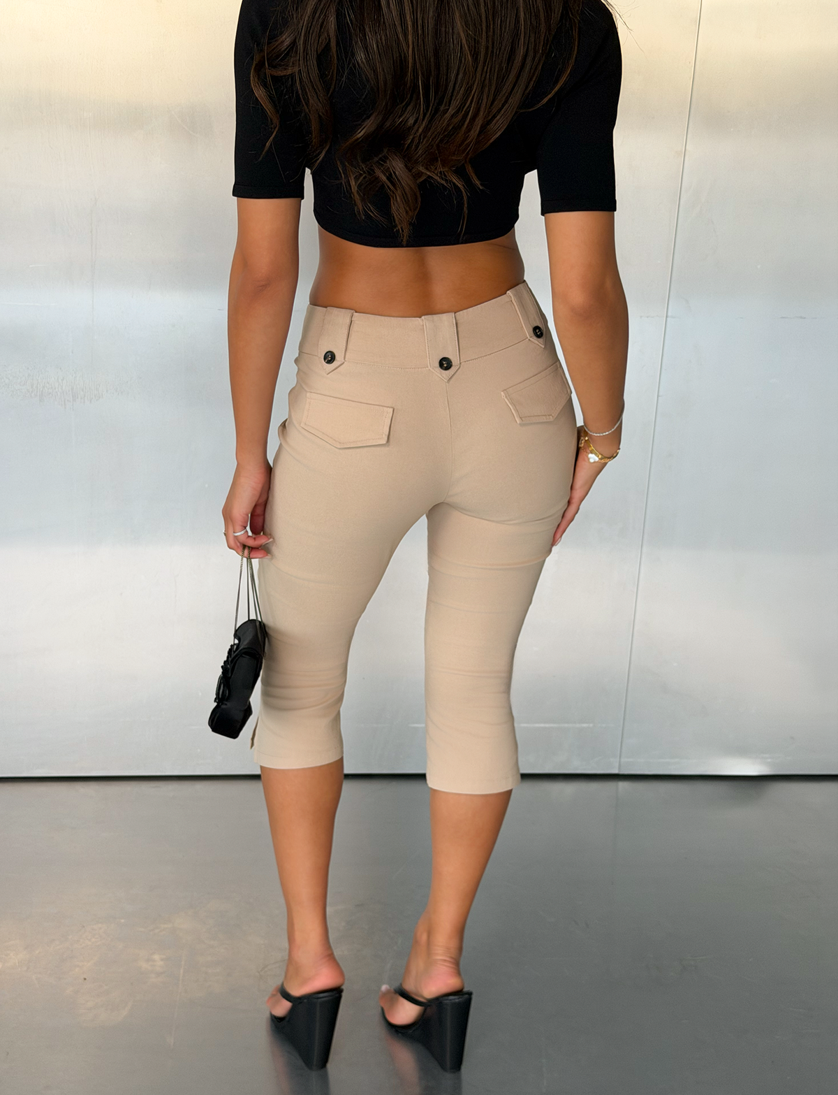 A woman stands indoors facing away, wearing a black crop top, the TIGERMIST ARIA CAPRI pants in khaki, black wedge heels, and holding a small black handbag. She has long brown hair.