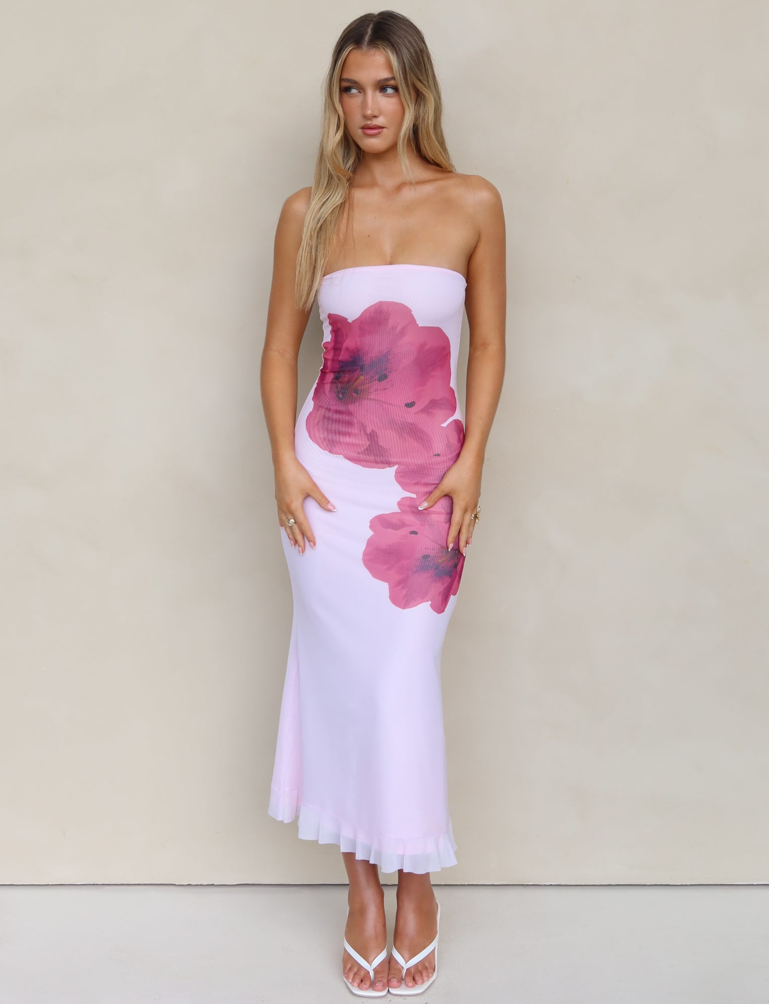 A woman with long blonde hair is wearing the LILLIAN DRESS - PINK by TIGERMIST, a strapless white dress adorned with large pink floral designs. She stands against a plain, beige background, complemented by her white, open-toed high heels. Her arms hang by her side as she gazes slightly downward, showcasing the elegant midi length of her outfit.