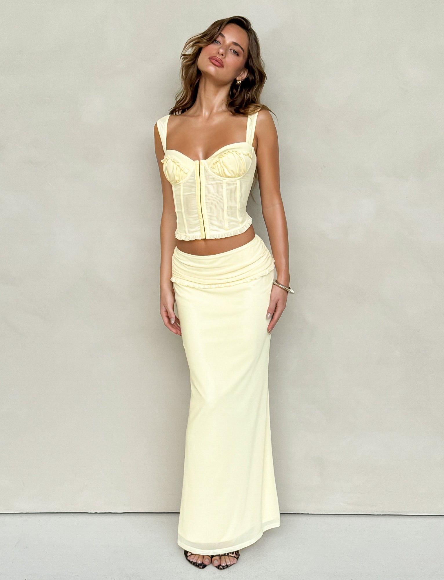A woman is posing against a plain backdrop, wearing a fitted yellow corset top and the TIGERMIST GINNY MAXI SKIRT - LEMON, which features a wide waistband. She stands with her head tilted and her hair flowing freely.