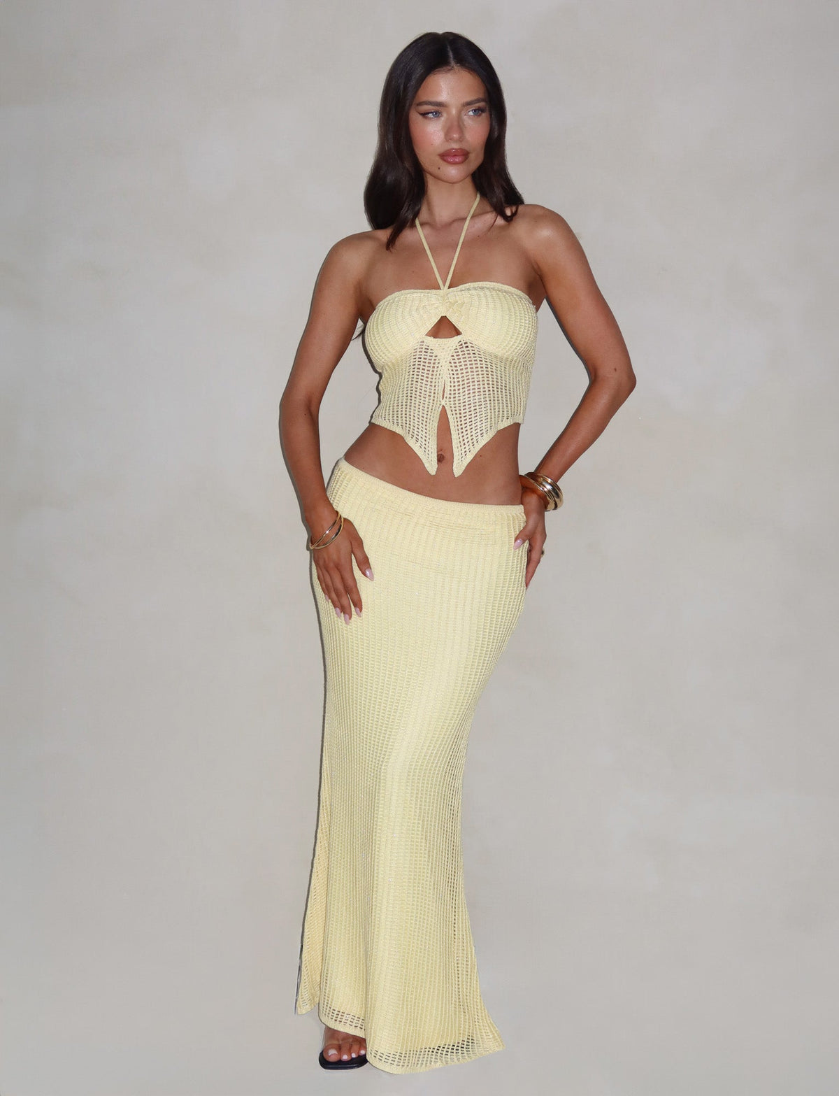 A woman with long dark hair is posing against a plain background, confidently showcasing the TIGERMIST SHAH SKIRT in yellow lemon. Her light yellow, form-fitting crochet two-piece outfit includes a halter top with intricate front details and a stylish maxi length skirt. She accentuates her look with bracelets and rings.