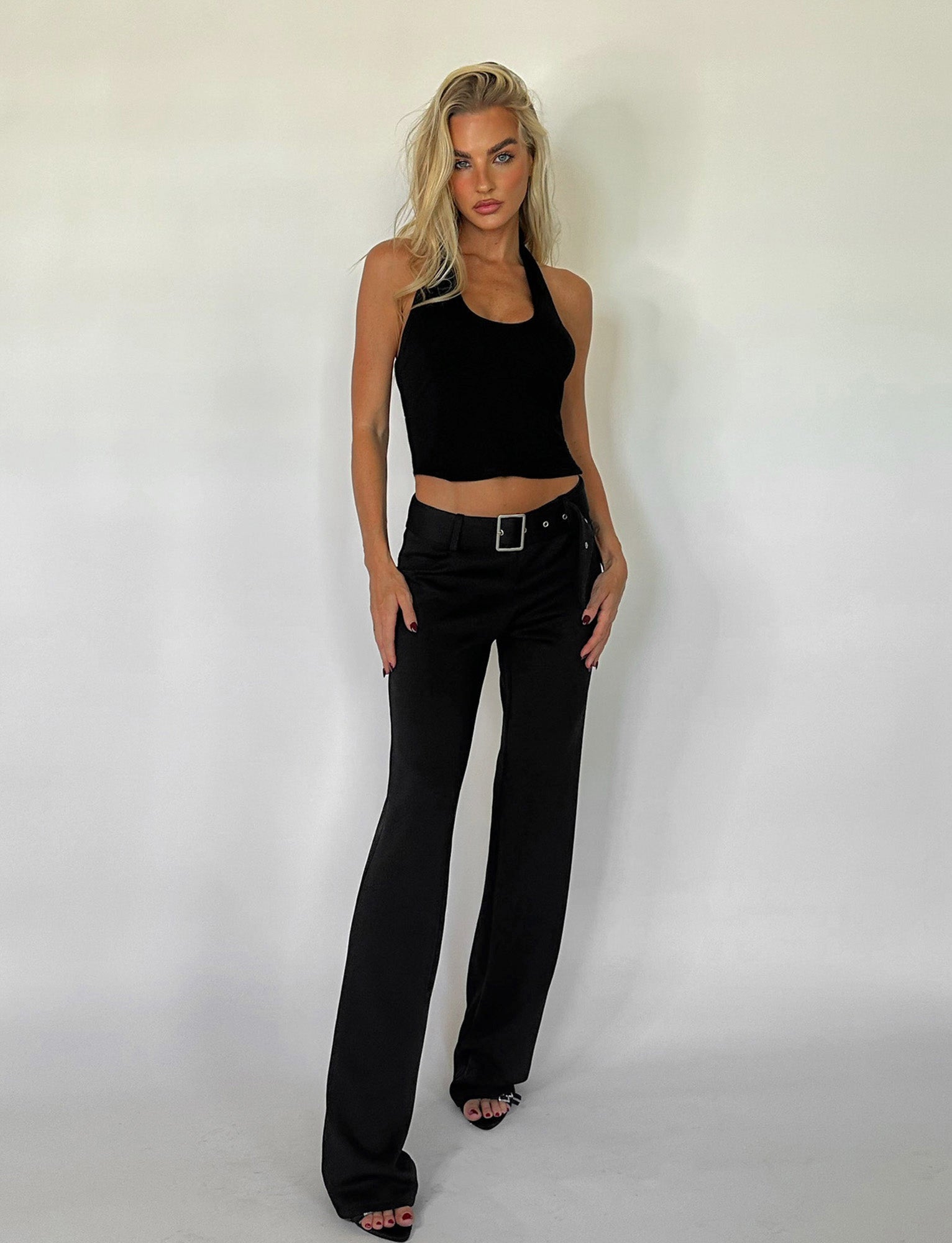 A person with long blonde hair stands confidently against a plain background, wearing a black sleeveless top and the HADLEY PANT - BLACK from TIGERMIST. They pose with one hand resting on their hip and the other lightly touching the side of their pants.