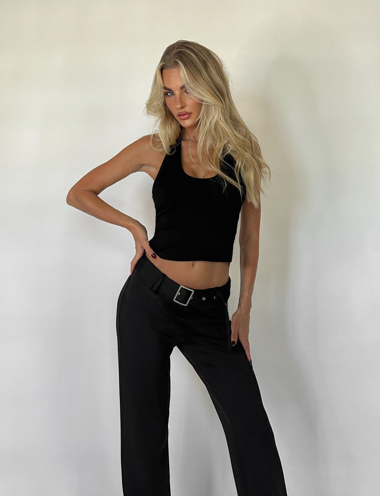 A person with long blonde hair wearing the ALEEN TOP - BLACK and black pants from TIGERMIST poses confidently against a plain white background, with one hand on their hip and a relaxed expression.