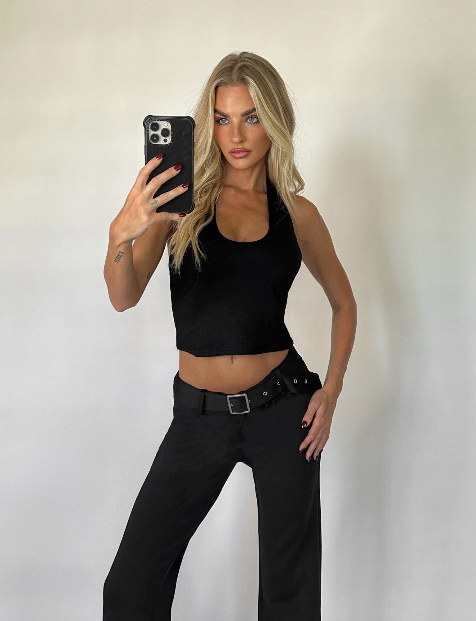 A person with long blonde hair is taking a mirror selfie with a phone. They are wearing the sleeveless ALEEN TOP - BLACK and black pants with a belt, both from TIGERMIST. The background is plain and neutral.