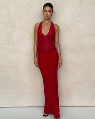 A woman poses against a textured beige wall, wearing the TIGERMIST BRESILY MAXI DRESS in red. The sleeveless dress features halter straps, and her straight dark hair is pulled back as she maintains a neutral expression.