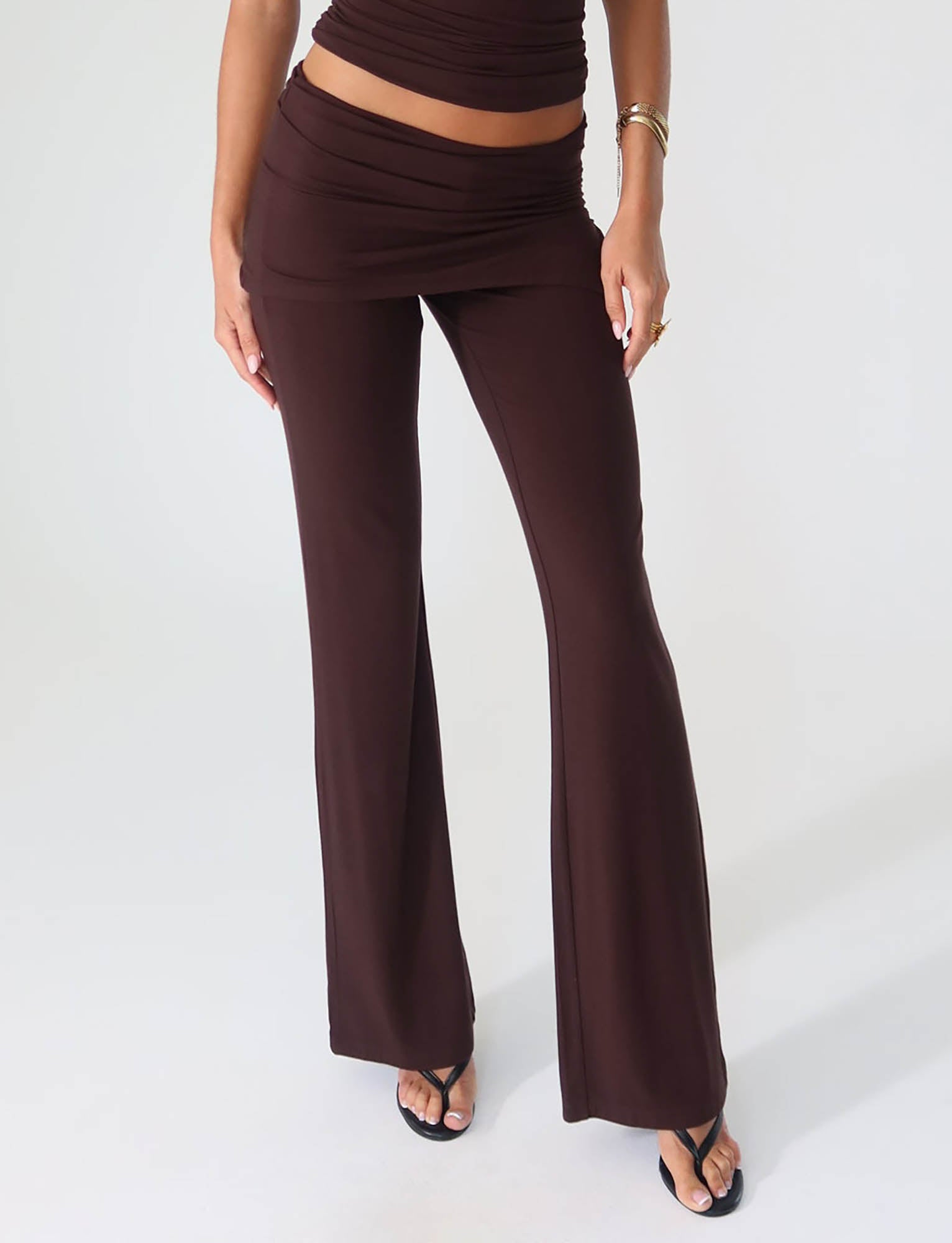 Against a plain white background, an individual showcases the LEILANI PANT - BROWN : CHOCOLATE from TIGERMIST, paired with a matching top that features a bare midriff. Crafted from high-stretch material for enhanced comfort, the ensemble is stylishly complemented by black sandals and gold jewelry.