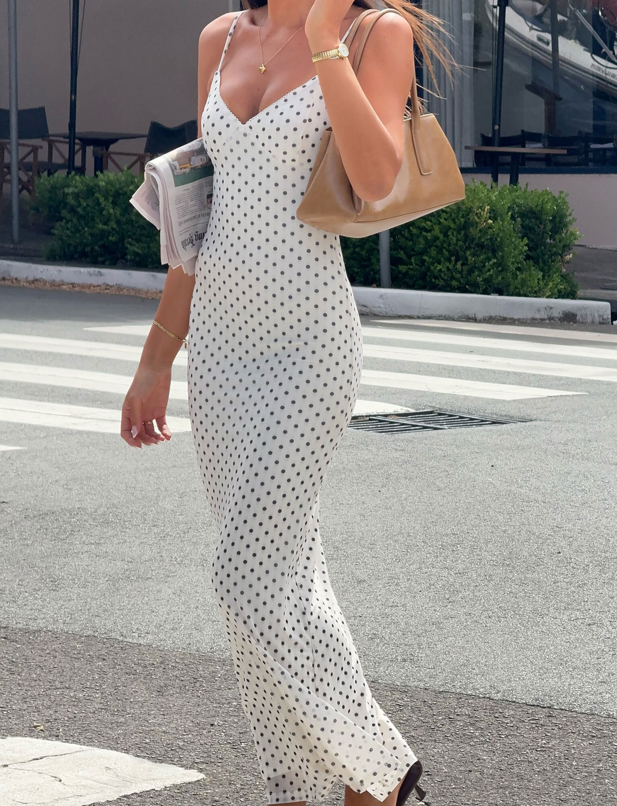 A woman wearing the TIGERMIST JULIANNA MAXI DRESS - POLKA DOT with adjustable straps walks on a city sidewalk, carrying a newspaper and tan handbag. She accessorizes with gold jewelry, brown sandals, and greenery with outdoor seating is in the background.