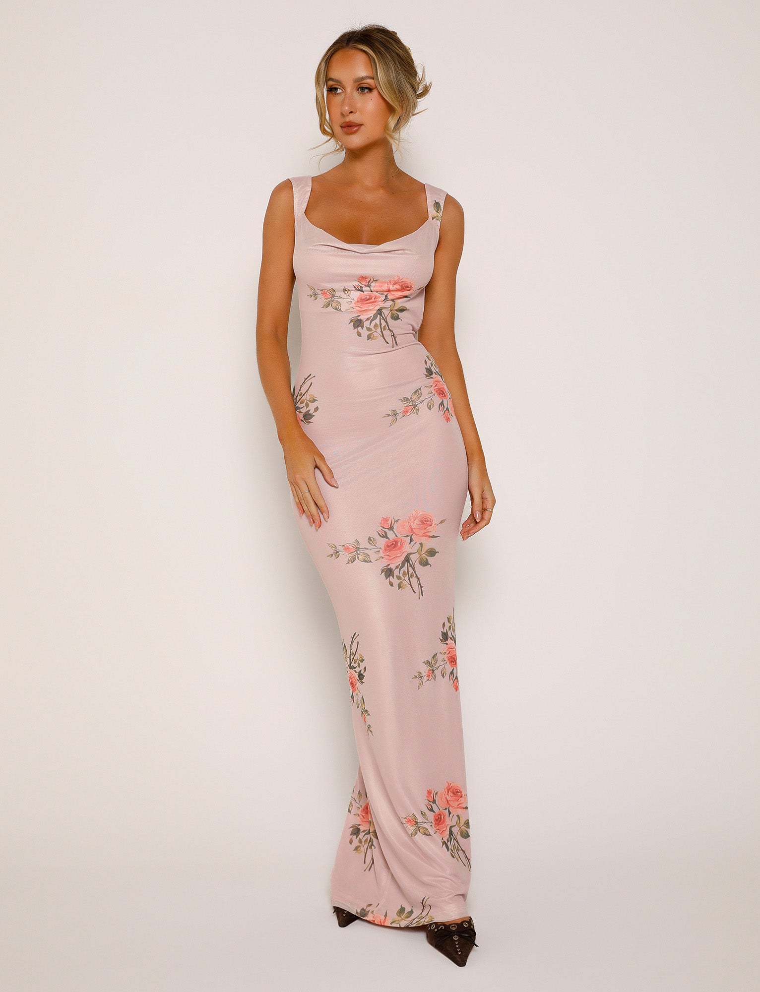 A person wearing the MORANA DRESS - PINK : FLORAL from TIGERMIST stands against a plain background. The long, form-fitting dress features a sleeveless design and a scoop neckline with elegant floral patterns. The individual has their hair styled in an updo and is looking slightly off to the side.