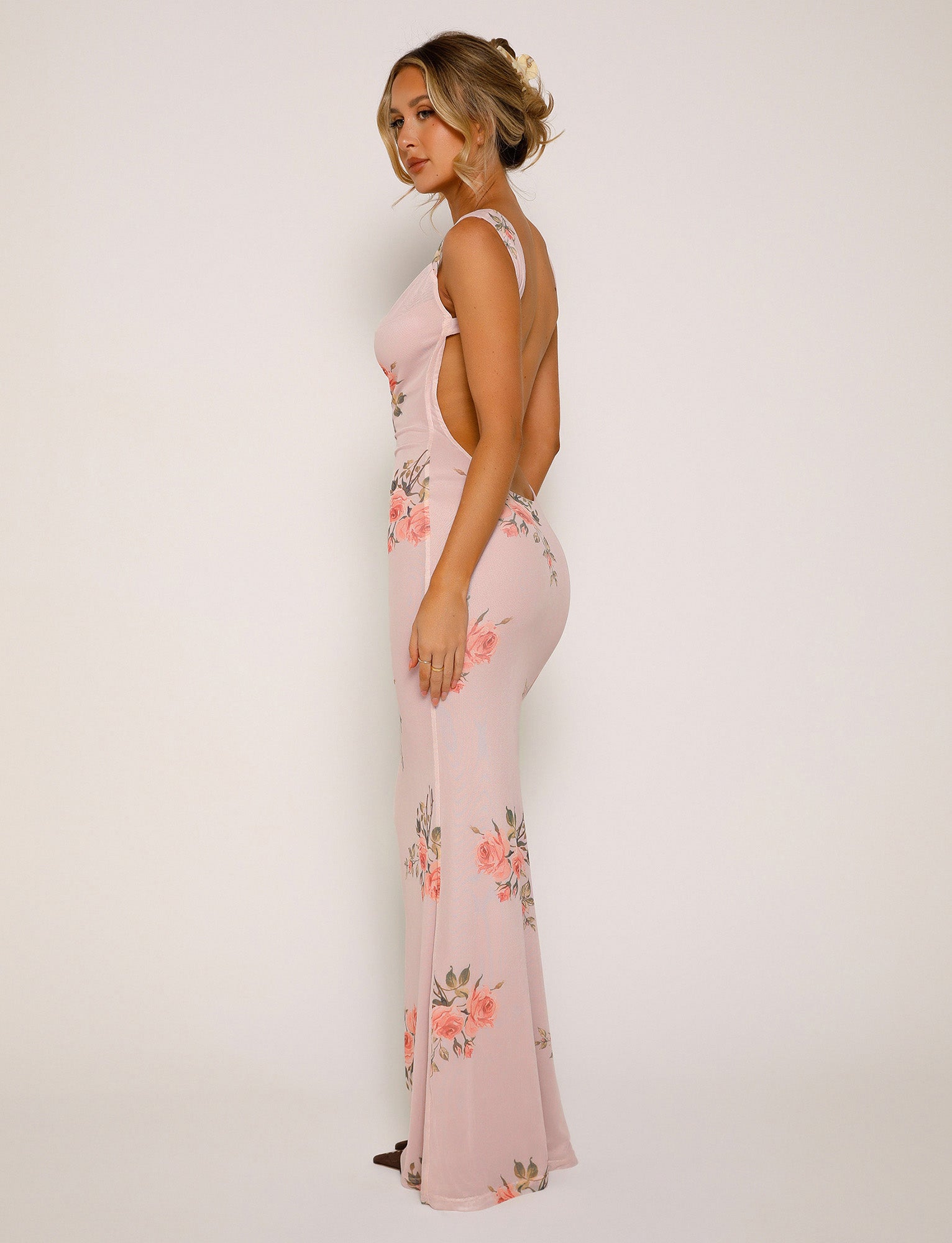 A woman stands against a plain background, wearing the MORANA DRESS - PINK : FLORAL from TIGERMIST. The long, form-fitting dress is adorned with floral patterns and features a low back and sleeveless design. She has blonde hair styled in an updo and is looking at the camera with a calm expression.