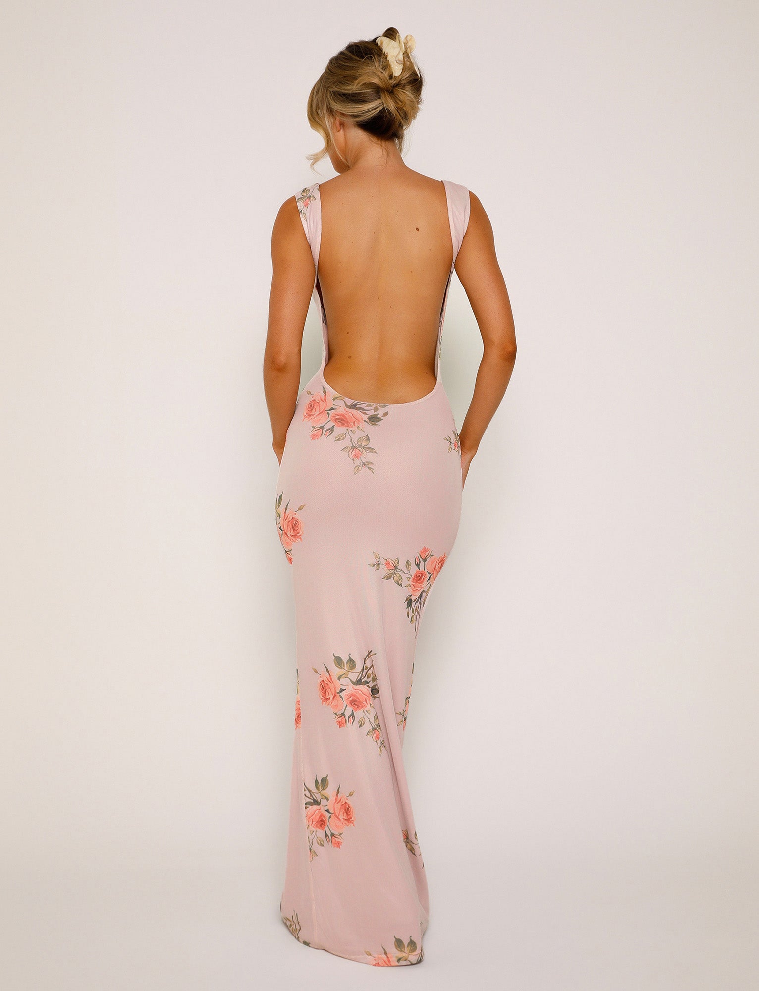 A woman is seen from the back wearing the MORANA DRESS - PINK: FLORAL by TIGERMIST, a long, light pink dress with a floral design. The dress features a deep open back and thick shoulder straps. Her hair is styled in a loose updo as she stands against a plain off-white background.