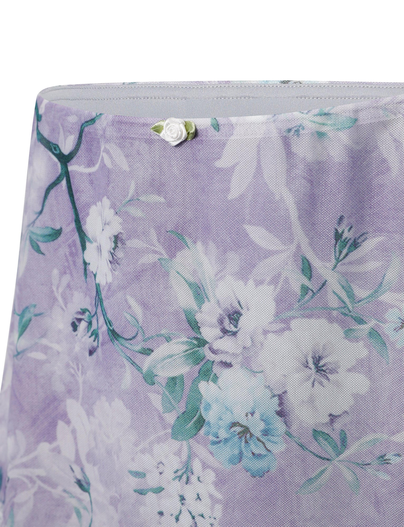 The AMARAN MINI SKIRT from TIGERMIST in a lavender hue features a floral pattern with white and light blue flowers and green leaves. A small white fabric rose decorates the top edge.