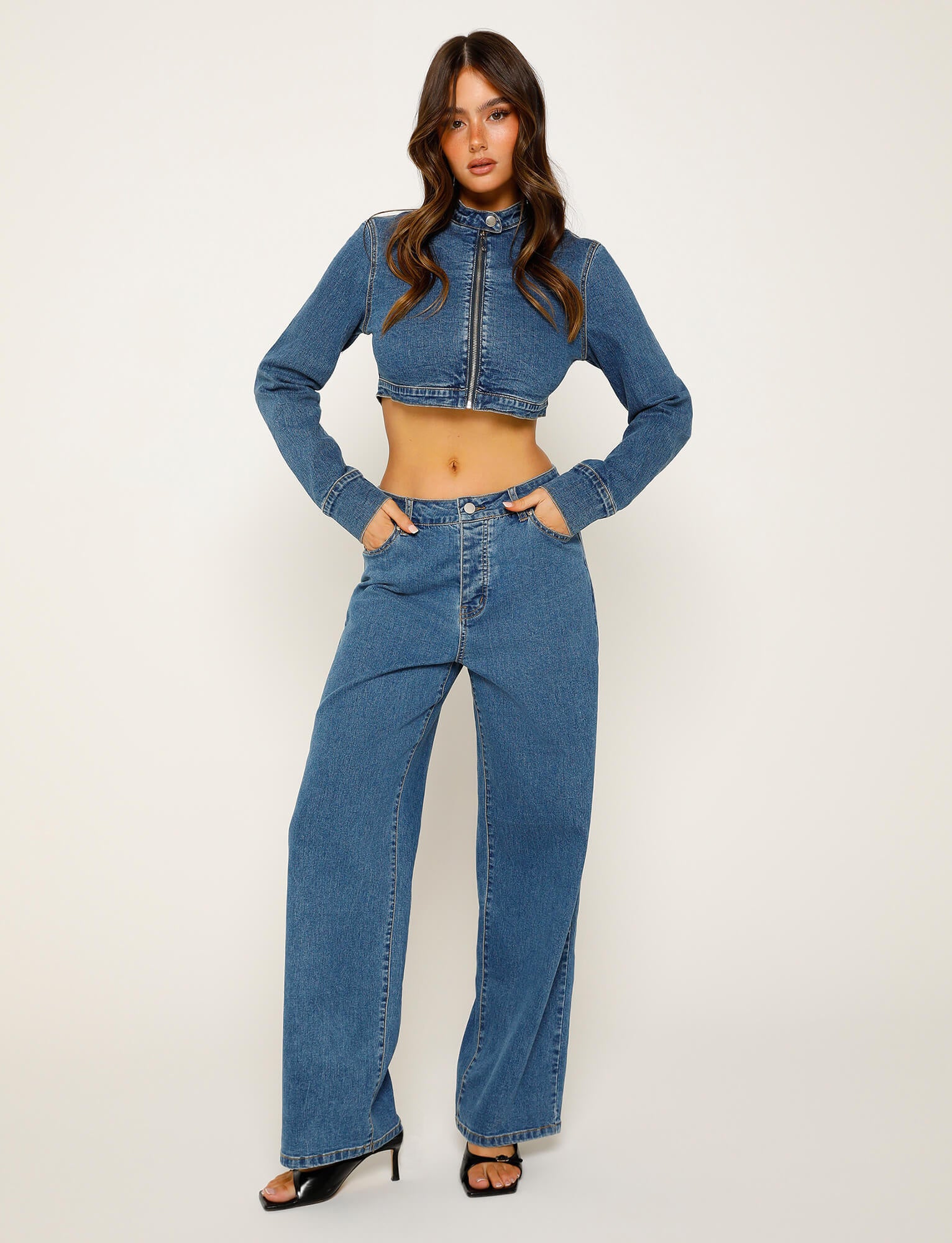 A young woman stands posing in a trendy TIGERMIST outfit. She wears the FELIX JEAN - BLUE : DARK WASH, consisting of a long-sleeve cropped denim jacket paired with high-waisted wide-leg denim pants. She has long wavy hair, with her hands in the front pockets of the pants, and she completes the look with black high-heeled sandals.