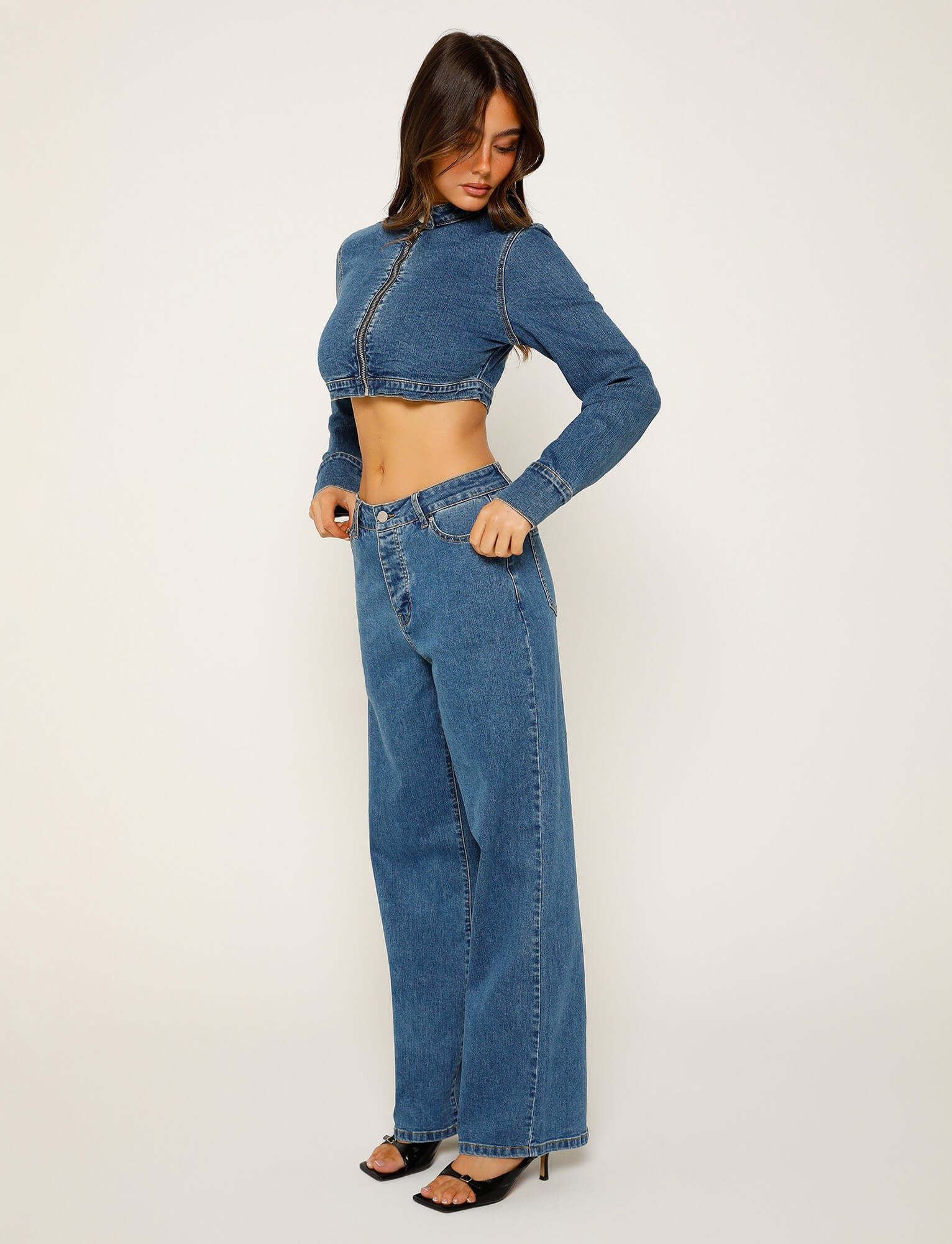 A woman poses against a plain background, wearing the FELIX JEAN - BLUE: DARK WASH from TIGERMIST. She is looking down and to the side, with her left hand resting in her pocket. She wears black, open-toed high heels.