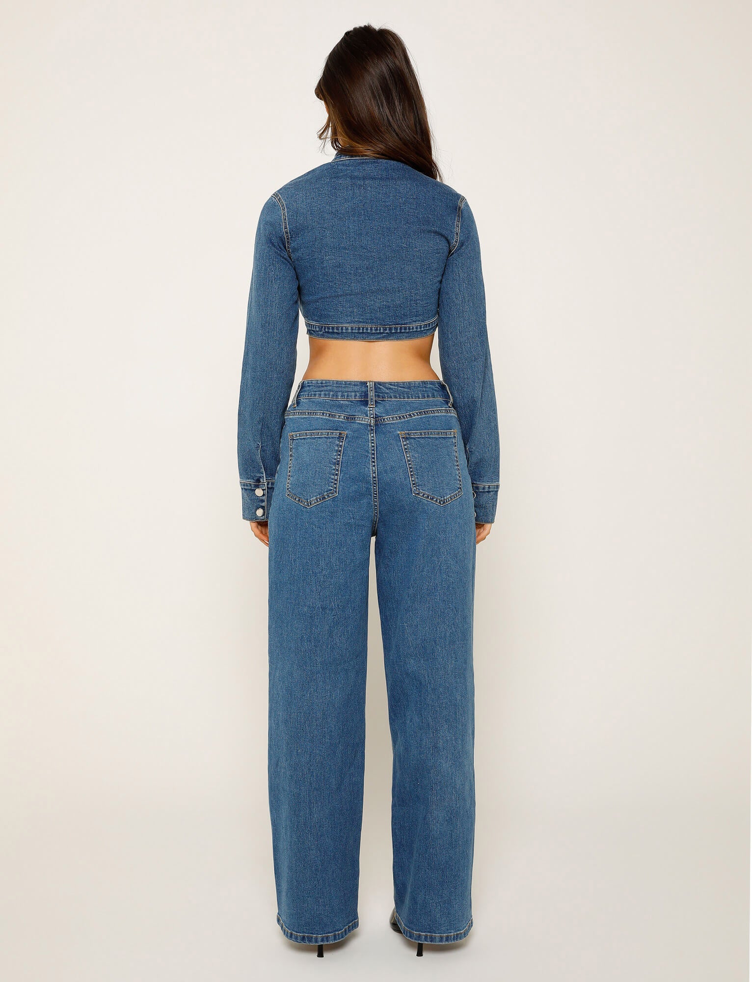 A person with long dark hair is standing with their back to the camera, wearing a long-sleeve, cropped denim jacket from TIGERMIST and high-waisted, wide-leg FELIX JEAN - BLUE : DARK WASH jeans. The background is plain and light-colored.