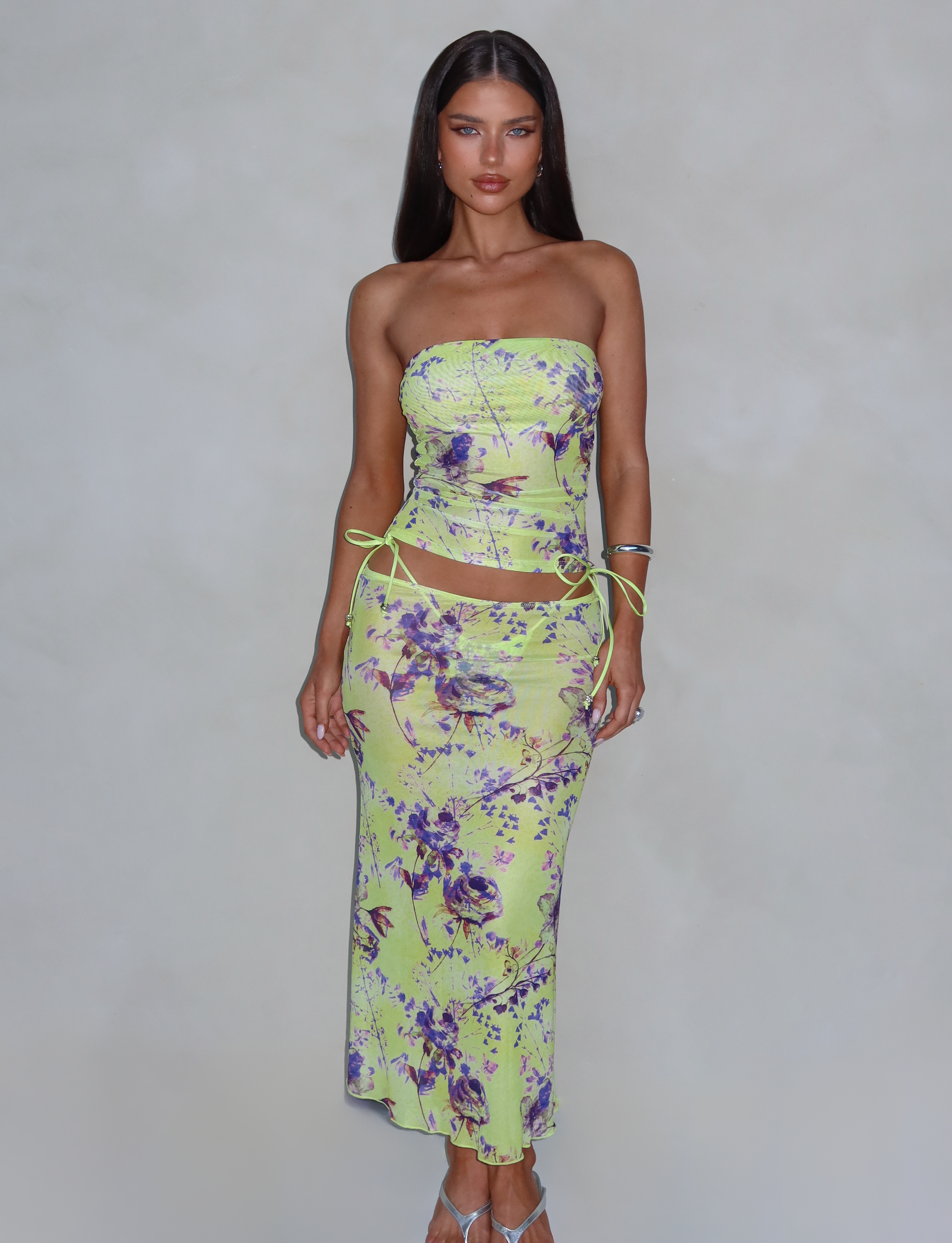 A woman with long dark hair poses against a plain background, wearing the KELLAN SKIRT - GREEN by TIGERMIST, featuring a strapless, floral print design. The yellow-green pattern adorned with purple flowers and sheer mesh detail elegantly flows to her knees as she stands with her hands by her sides, gazing directly at the camera.