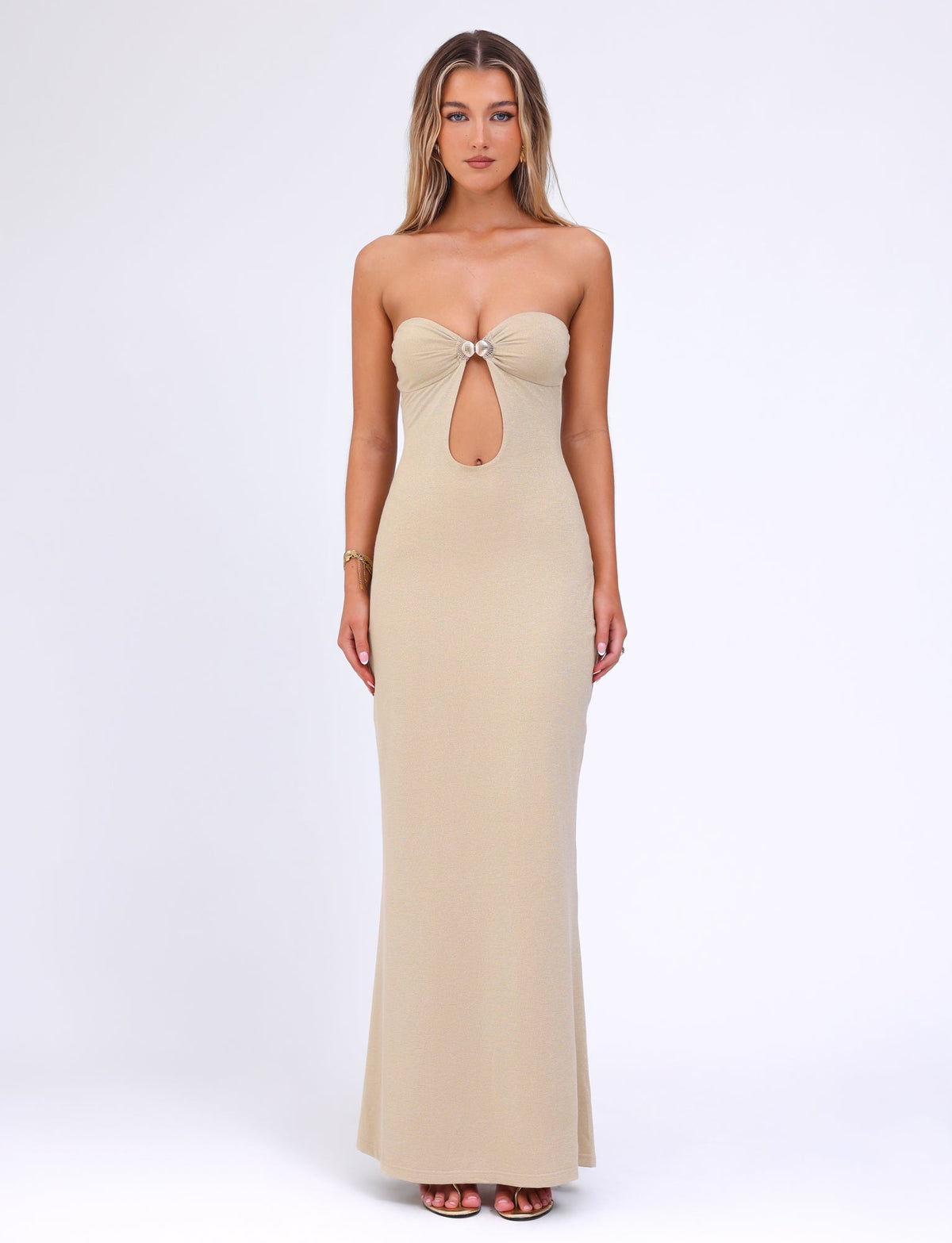 A woman stands posing in the CECILY MAXI DRESS - GOLD by TIGERMIST, a floor-length, fitted beige gown featuring a circular ring at the bust and a keyhole cutout below. The plain white background accentuates the dress's elegant design and its luxurious gold colourway.