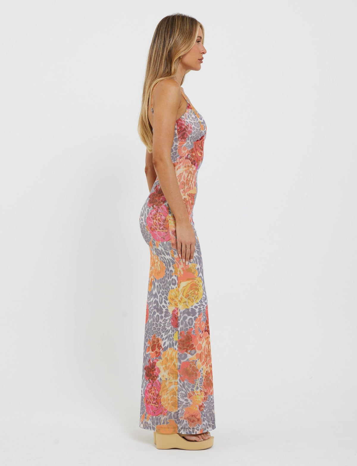 A woman stands in profile against a white background, wearing the TIGERMIST Julianna Dress - Multi: Leopard: Orange Leopard, which features a colorful floral and leopard print pattern. She has long, straight hair and is wearing platform shoes.