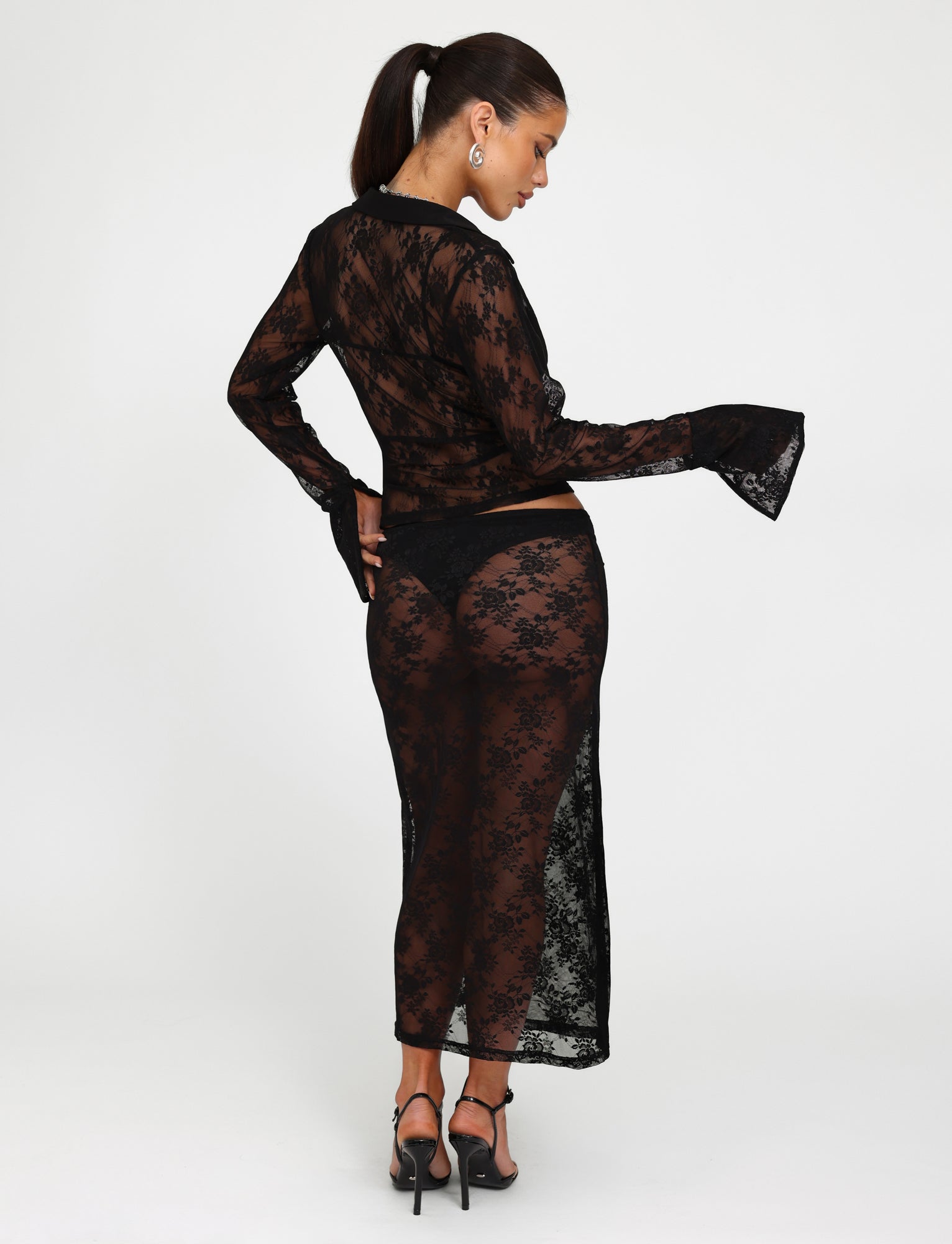 A woman stands in a studio, facing away from the camera, wearing a TIGERMIST black lace long-sleeve top and the matching MIRABEL SKIRT - BLACK. Her hair is tied in a ponytail and she is wearing black high-heeled sandals.