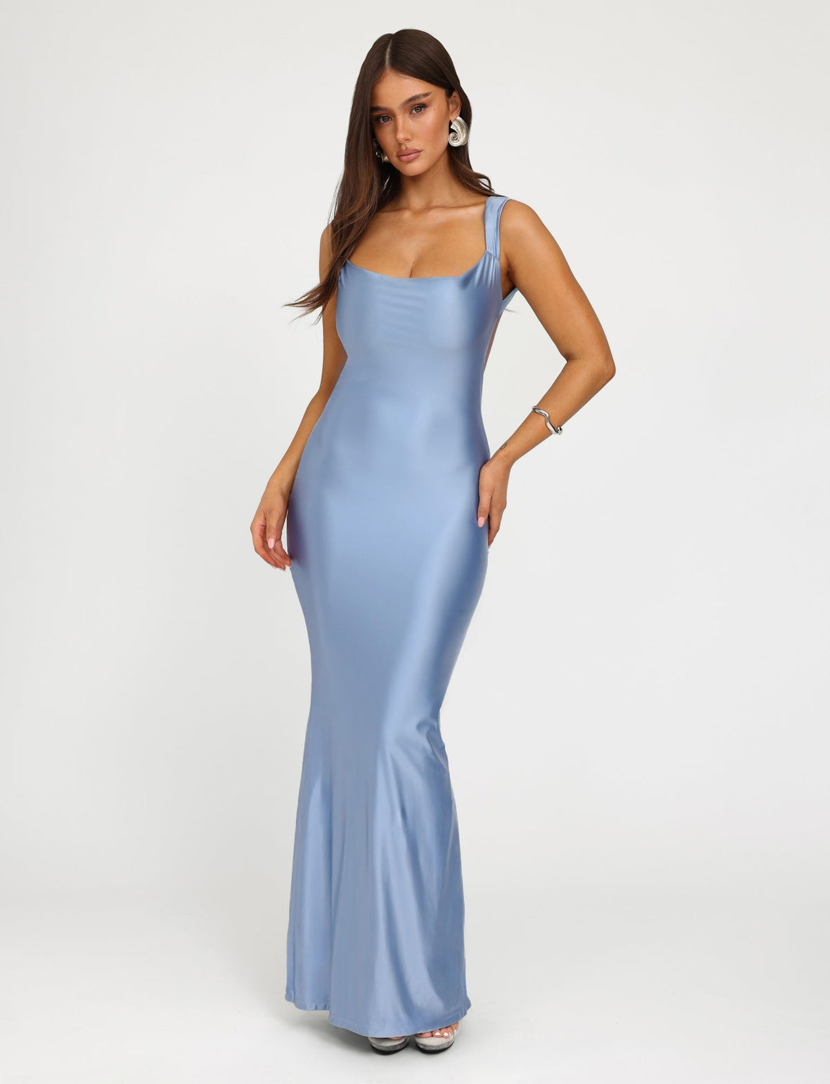 A woman is wearing a form-fitting, silky pale blue evening gown called the MORANA SHIMMER DRESS from TIGERMIST, featuring a scoop neckline. She has long hair and is posing with her left hand on her hip and her right arm relaxed. She is also wearing hoop earrings and a bracelet. The background is plain white.