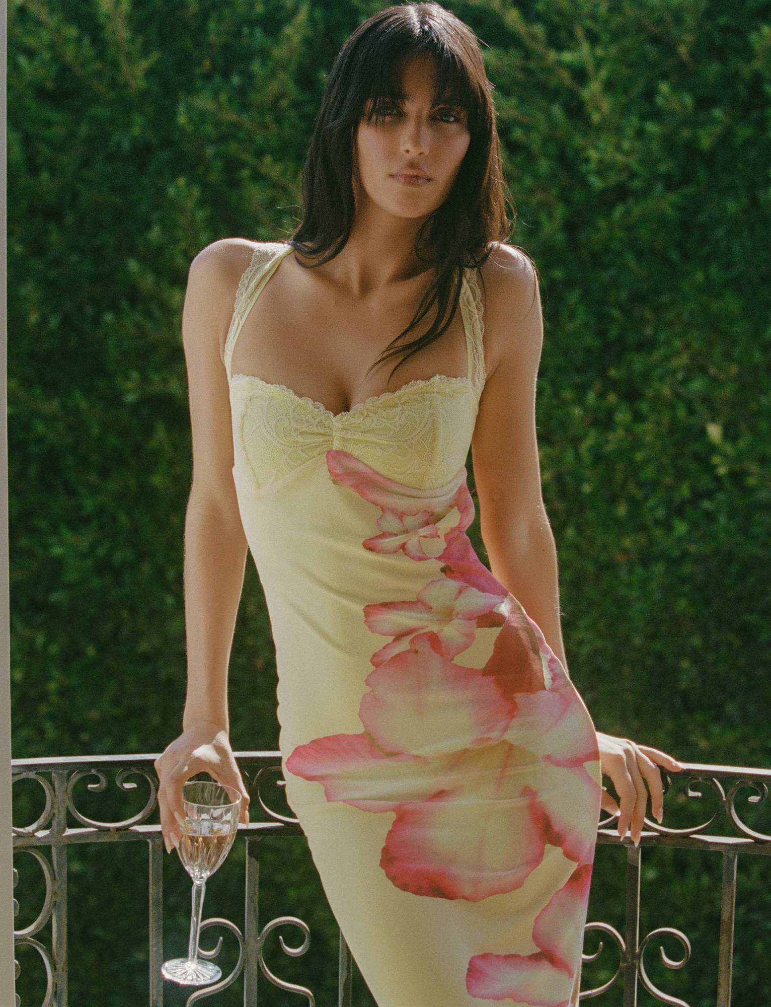 A woman with long dark hair stands outdoors by a metal railing, holding a glass of white wine and wearing the TIGERMIST NYOMI MAXI DRESS in yellow hibiscus macro floral print, surrounded by lush green foliage.