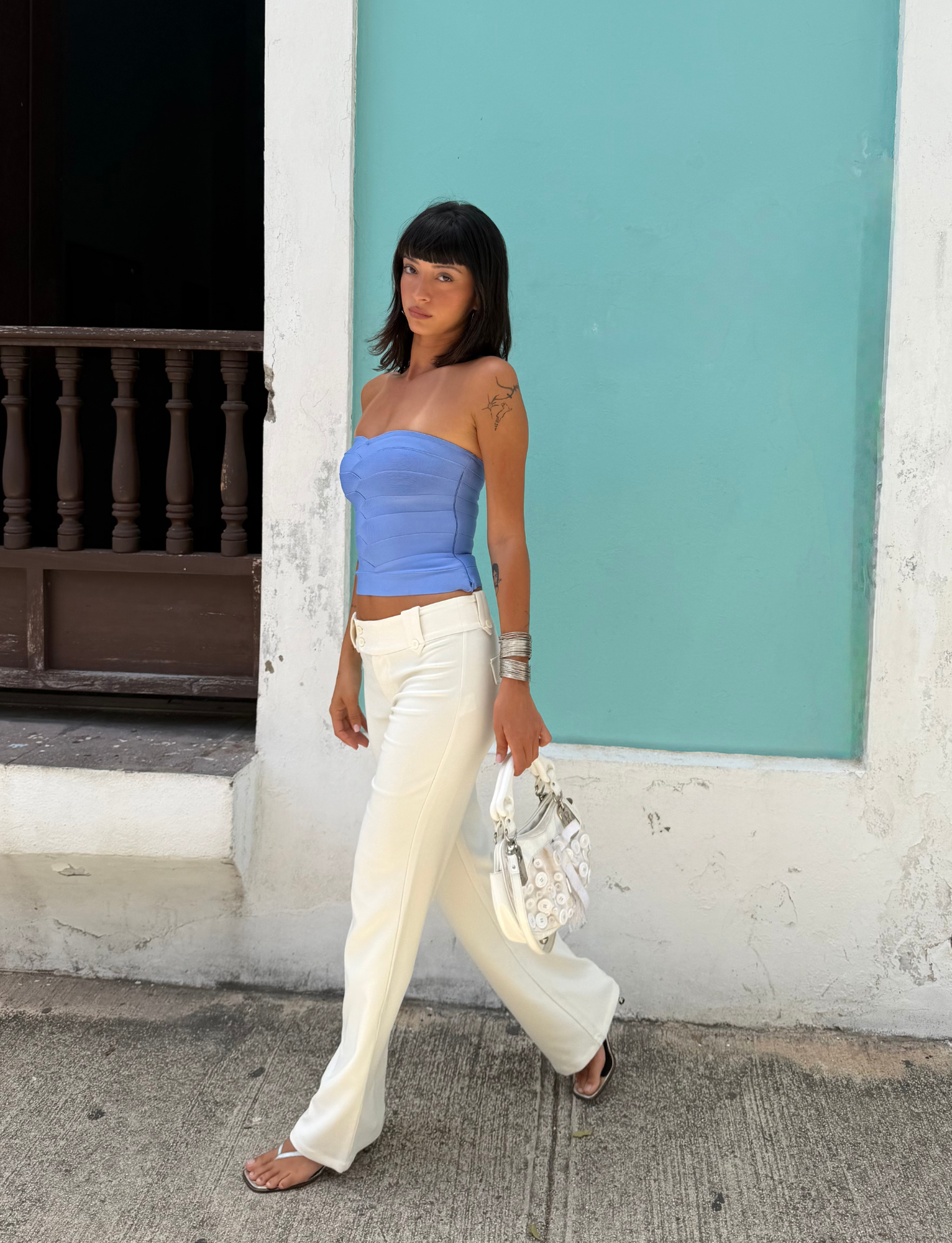 Wearing the TIGERMIST NIYA BANDAGE TOP in Cornflower Blue, a woman with dark hair pairs it with white pants as she walks past a turquoise wall, accessorizing with silver jewelry and carrying a white bag.