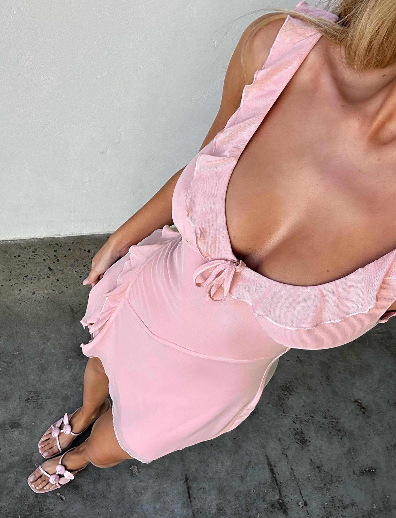 A person is wearing the TIGERMIST IMRA DRESS in dusty pink, featuring ruffled edges and a bow at the neckline. They are wearing sandals with bow accents and standing on a concrete surface. The image is taken from a high angle, focusing on their upper body and attire.