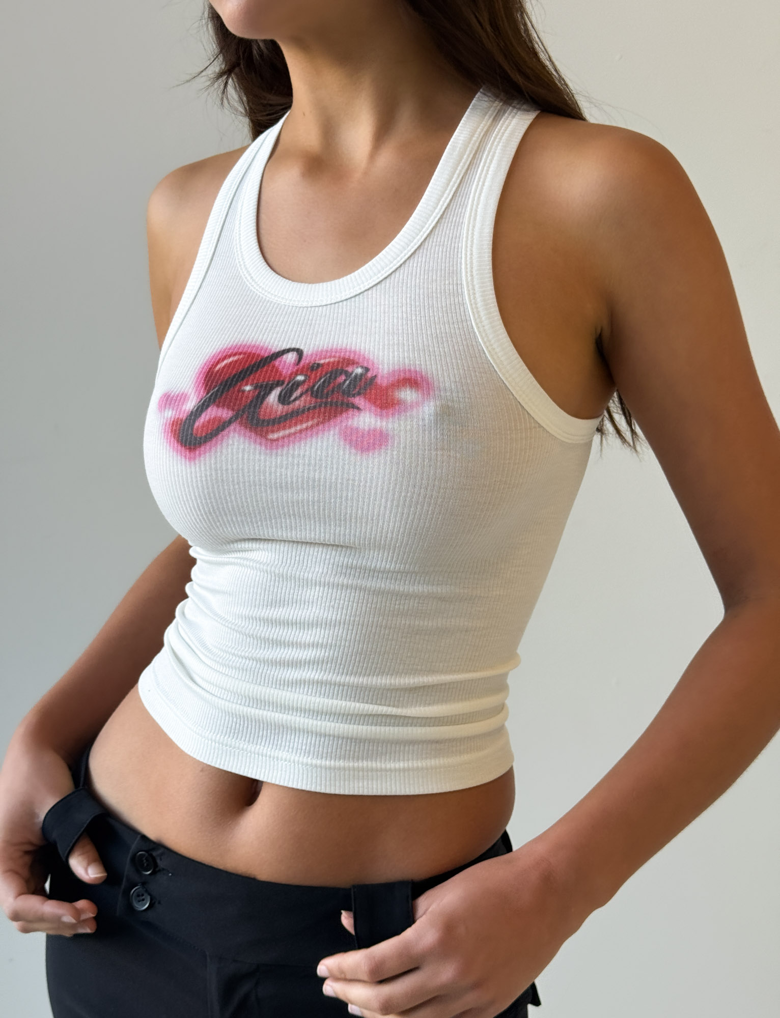 A woman wears the I.AM.GIA PENELOPE TANK in pink airbrush lettering, a white ribbed racer tank, with black pants against a plain light background.