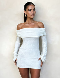 A woman with long dark hair in a sleek ponytail is wearing the Diedre Jumper Dress - White : Cream by TIGERMIST. She has gold hoop earrings and is posing against a beige background. With a serious expression, her hands rest relaxed near her hips.