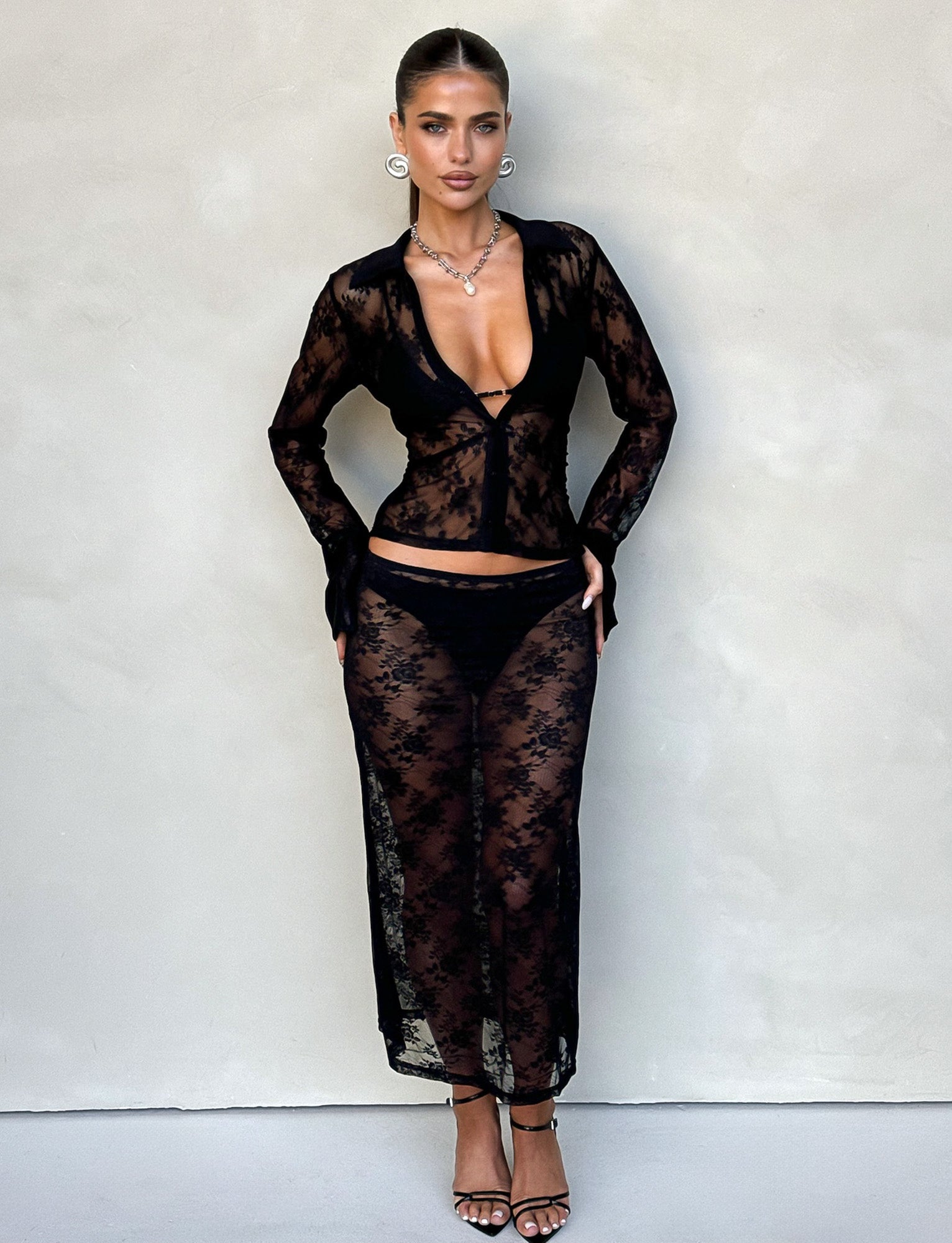 A woman stands against a beige wall wearing TIGERMIST's MIRABEL SKIRT in black lace, featuring a sheer long-sleeved top and skirt with visible black underwear beneath. She accessorizes with hoop earrings, a necklace, and black high-heeled sandals, with her hair styled in a sleek, pulled-back look.
