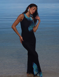 A woman in the TIGERMIST ZAHRAA DRESS - BLACK : BUTTERFLY stands in shallow water. She gently touches her wet hair, looking down with a serene expression. The calm sea in the background merges with the clear sky, creating a tranquil scene.